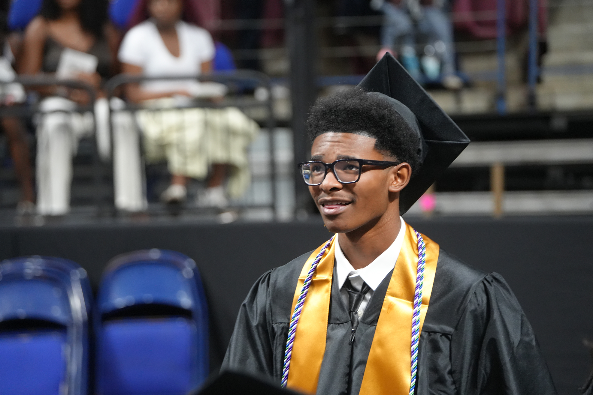 A young person in a black gown and cap stands smiling on a stage with empty chairs in the background.
