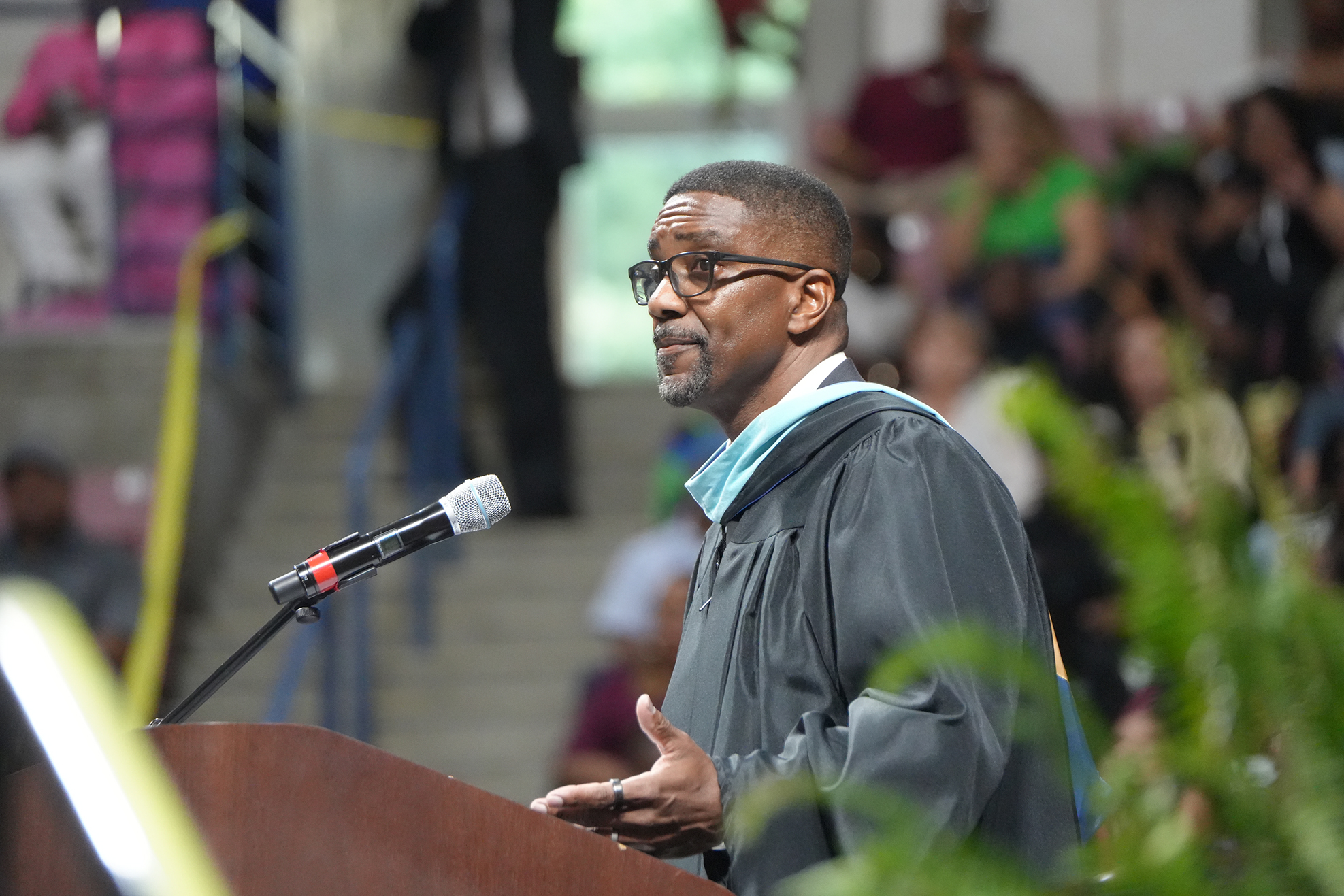 A man in a black academic gown speaks into a microphone at a podium, with an audience seated in the background.
