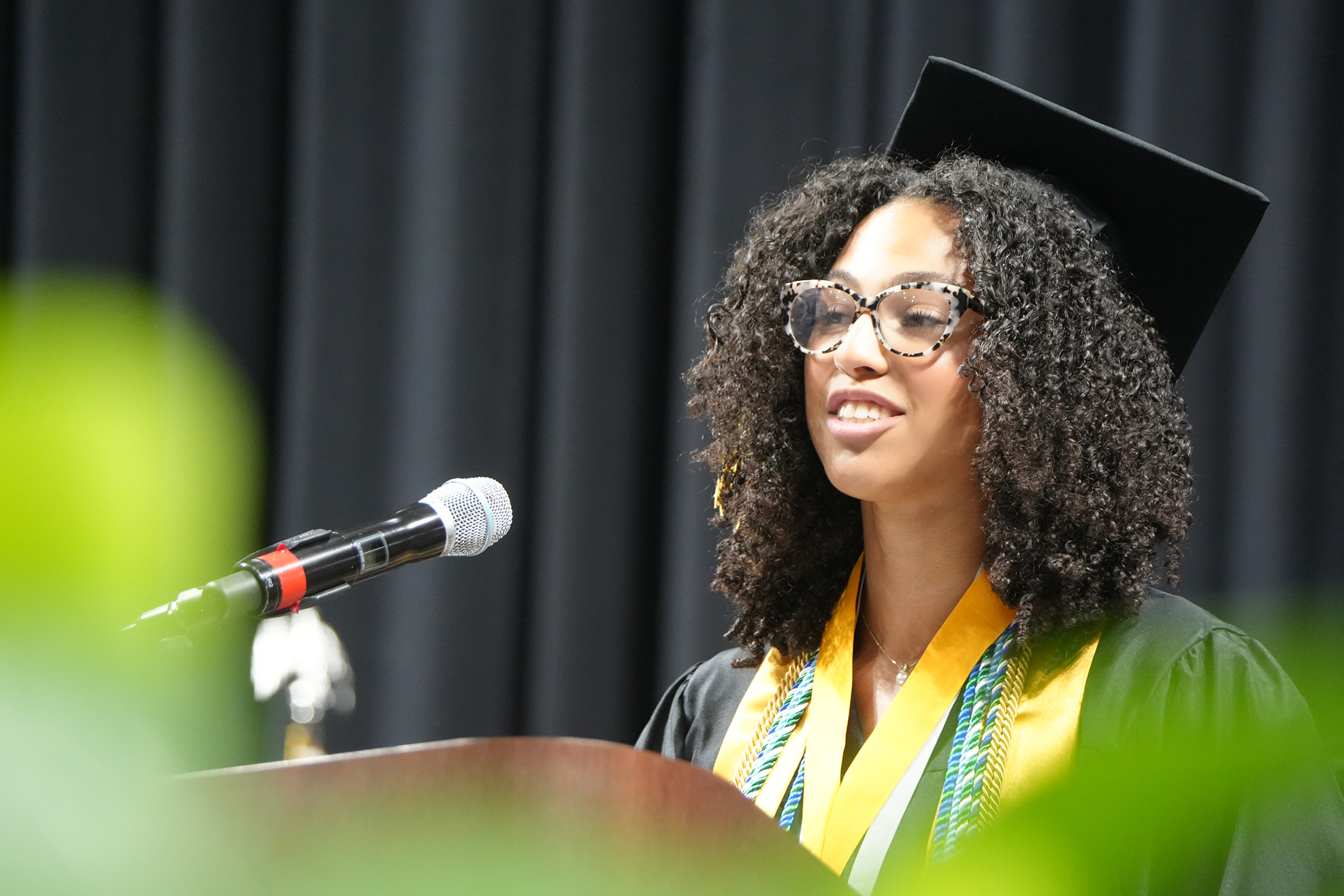 A woman with curly hair and glasses, wearing a graduation gown, speaks into a microphone at an event.