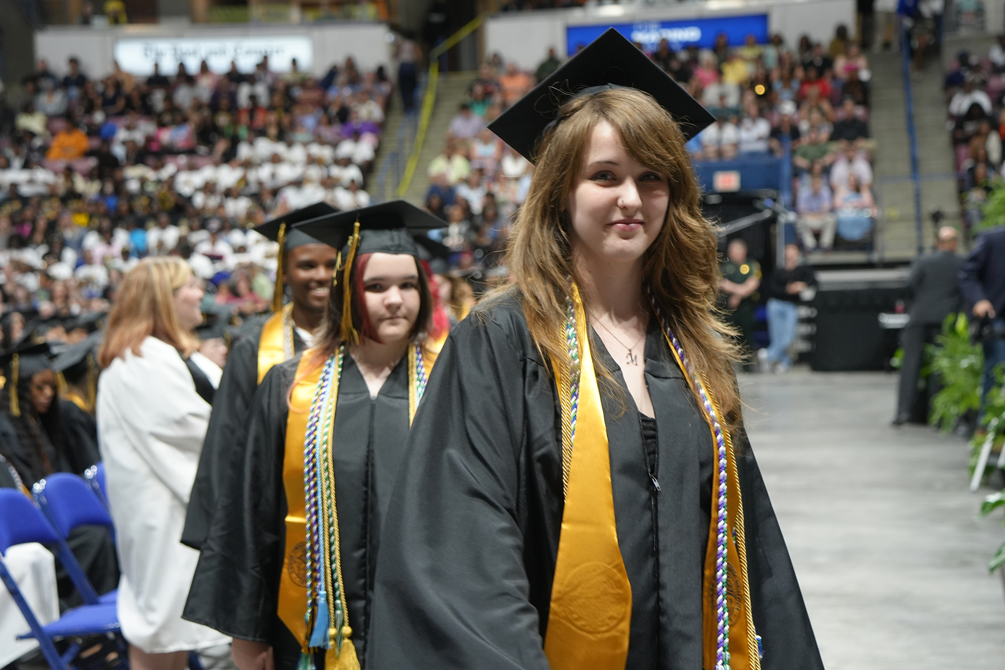 Three women in graduation gowns and caps walk on stage in an auditorium, with a crowd in the background.