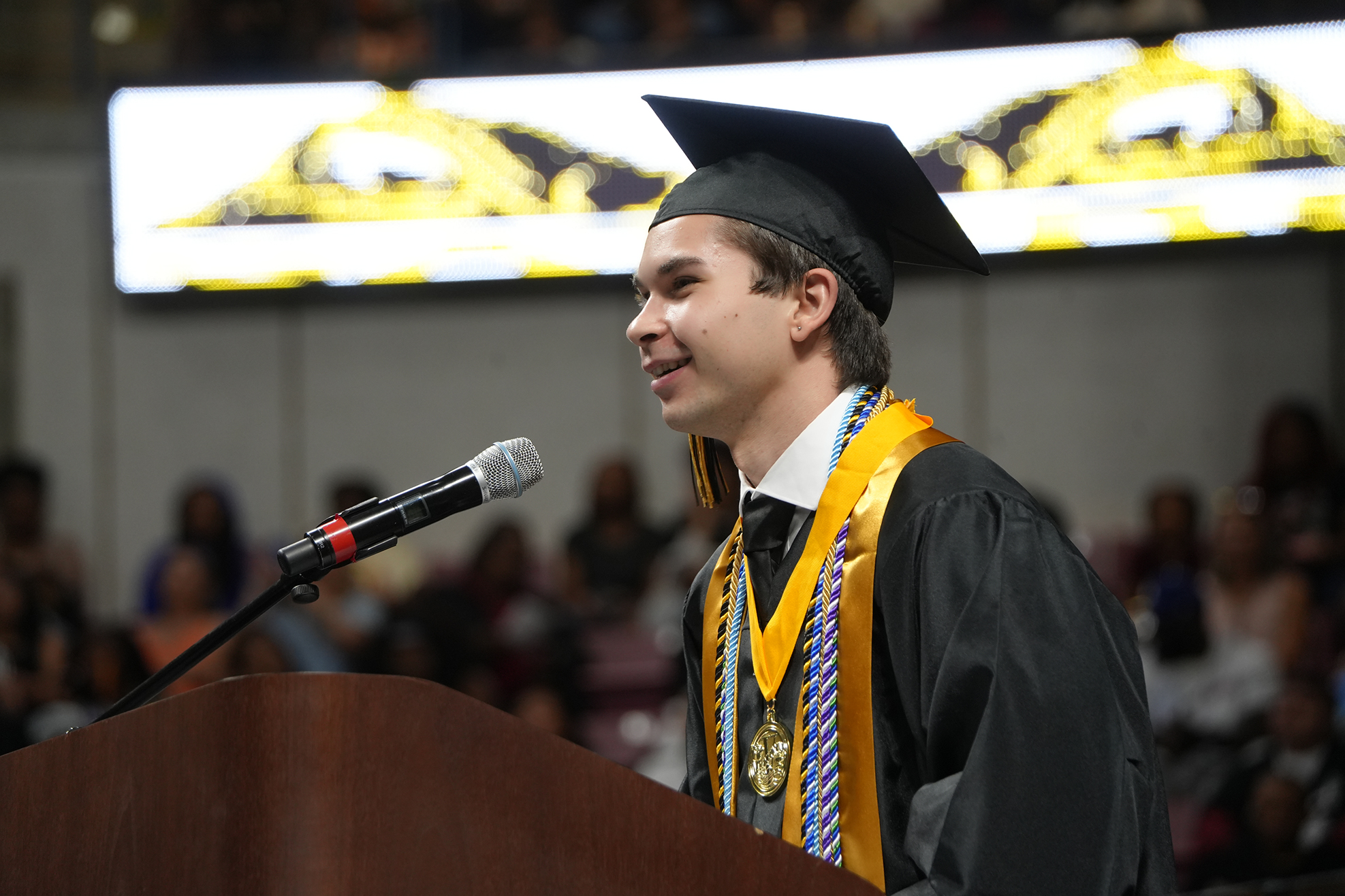 A young male in a graduation cap and gown standing at a podium with a microphone.