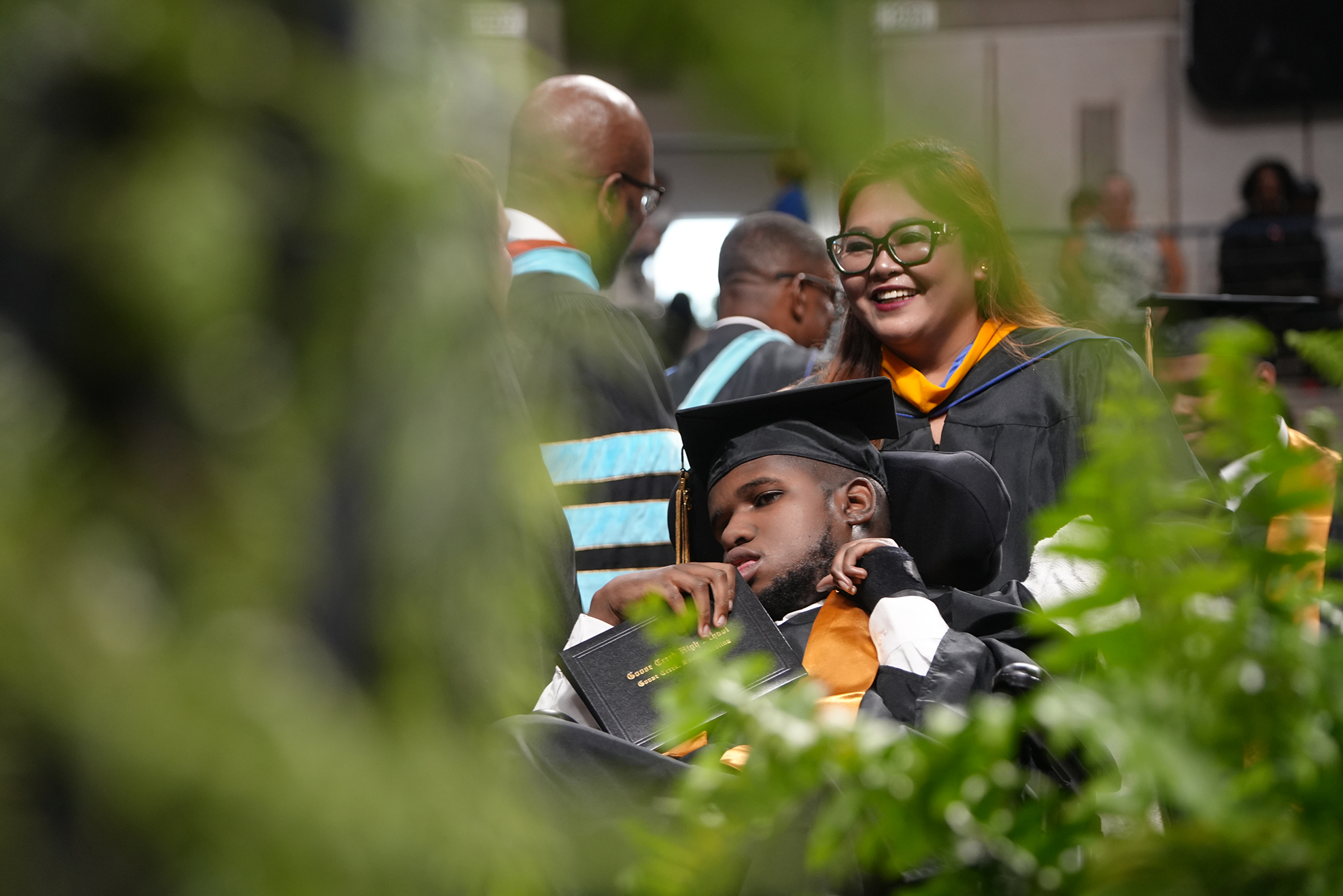 People at a graduation ceremony. A man in a wheelchair is smiling, with a diploma.