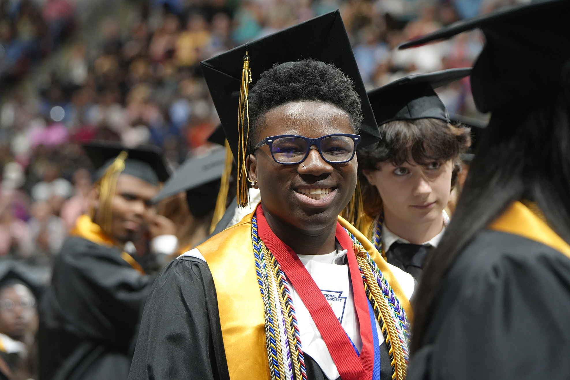 Several people in black graduation gowns, caps, and cords, standing in an audience. The central person smiles.