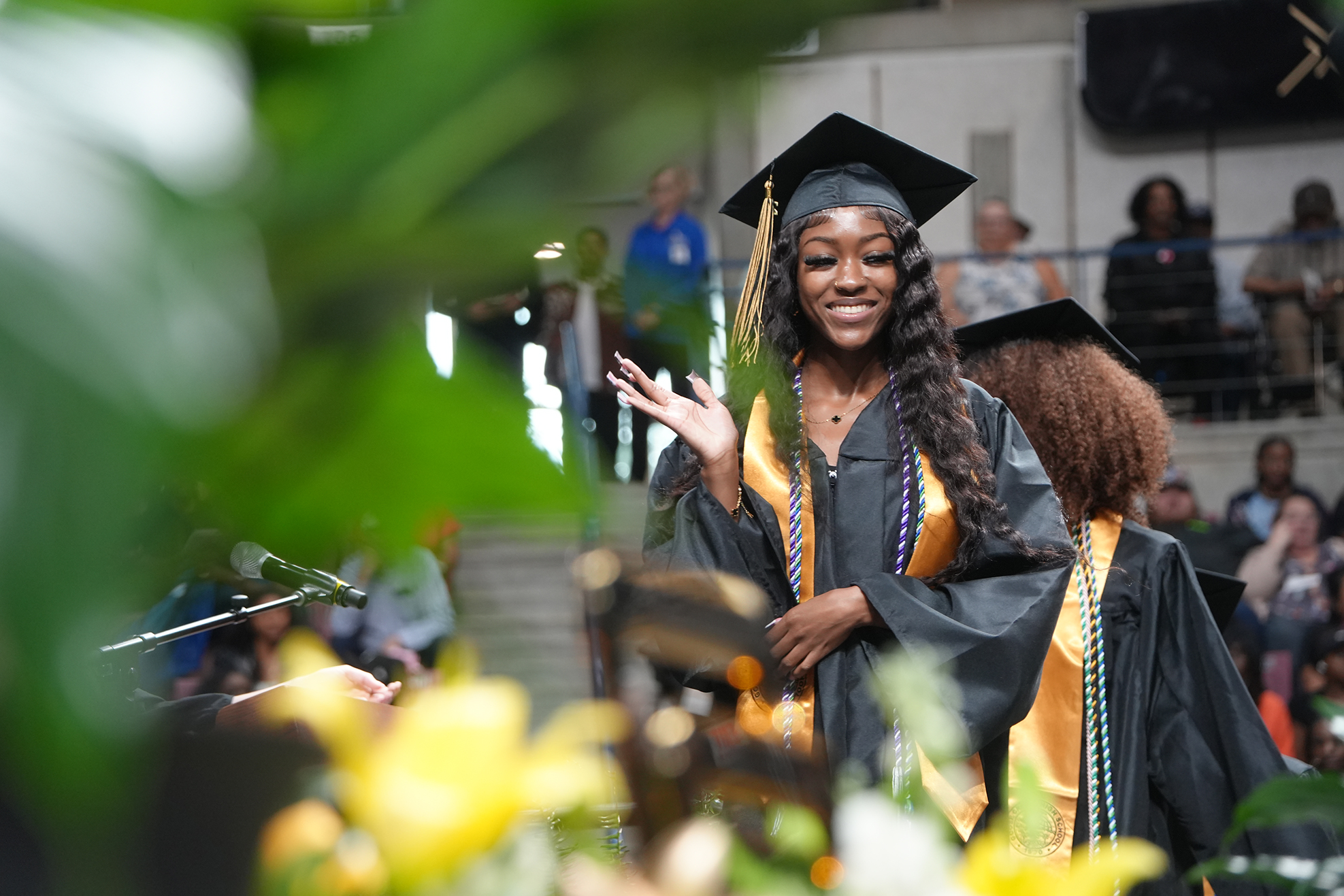 A woman in a black graduation gown waves, wearing a cap. Behind her, people stand on stairs, watching.