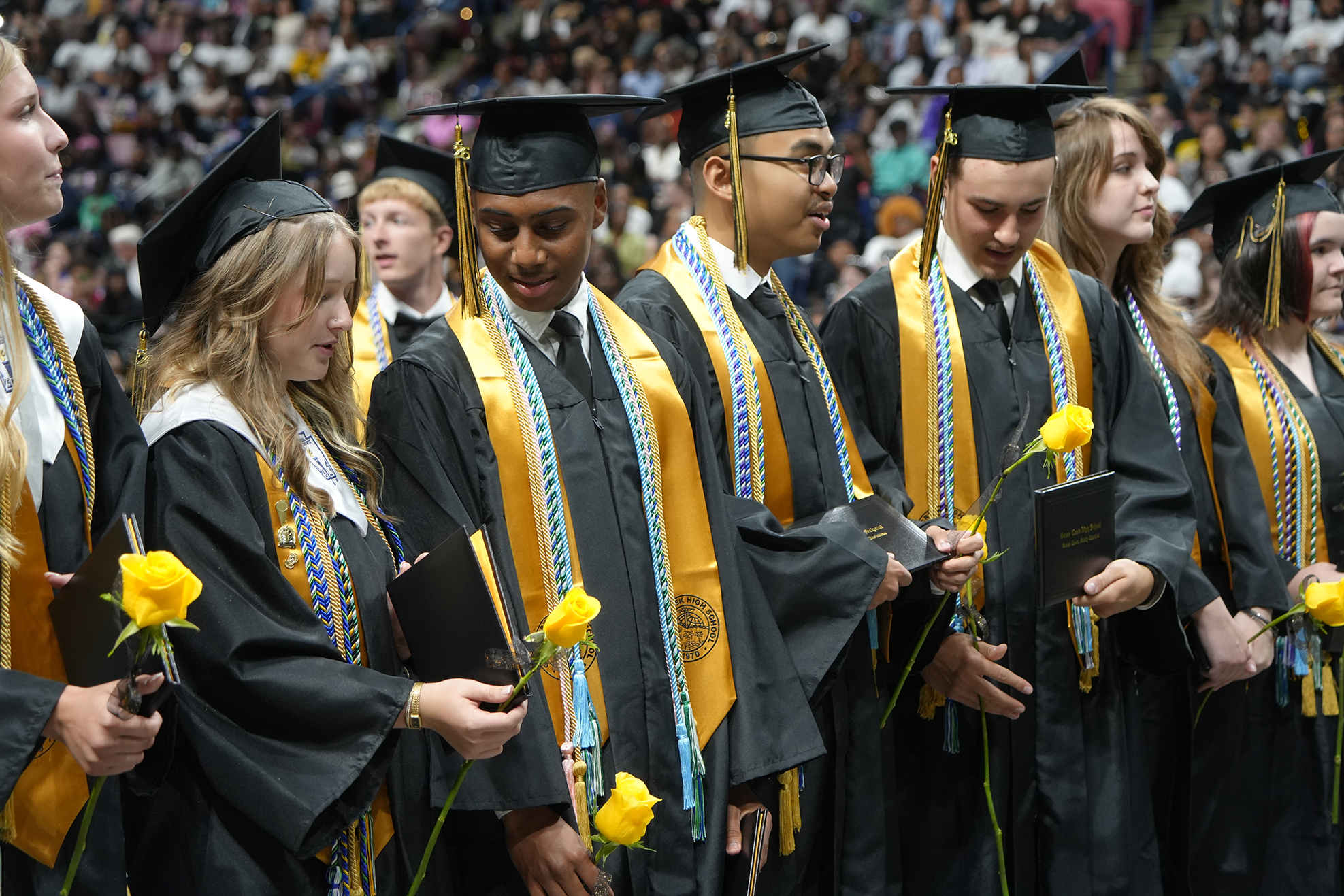 A row of students in graduation attire, holding yellow flowers, with a large audience in the background.