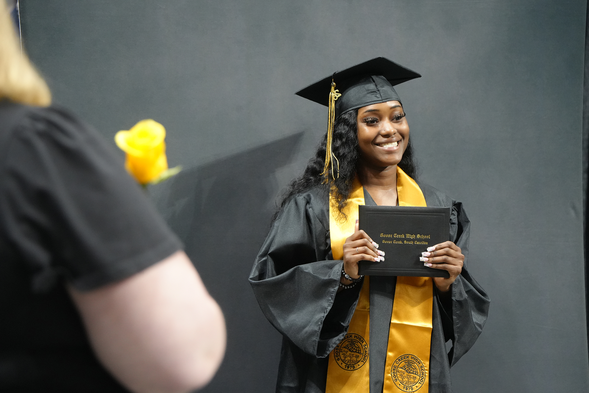 A woman in a graduation gown holds a diploma and smiles at the camera. Another person stands nearby, partially out of frame, holding a yellow flower.