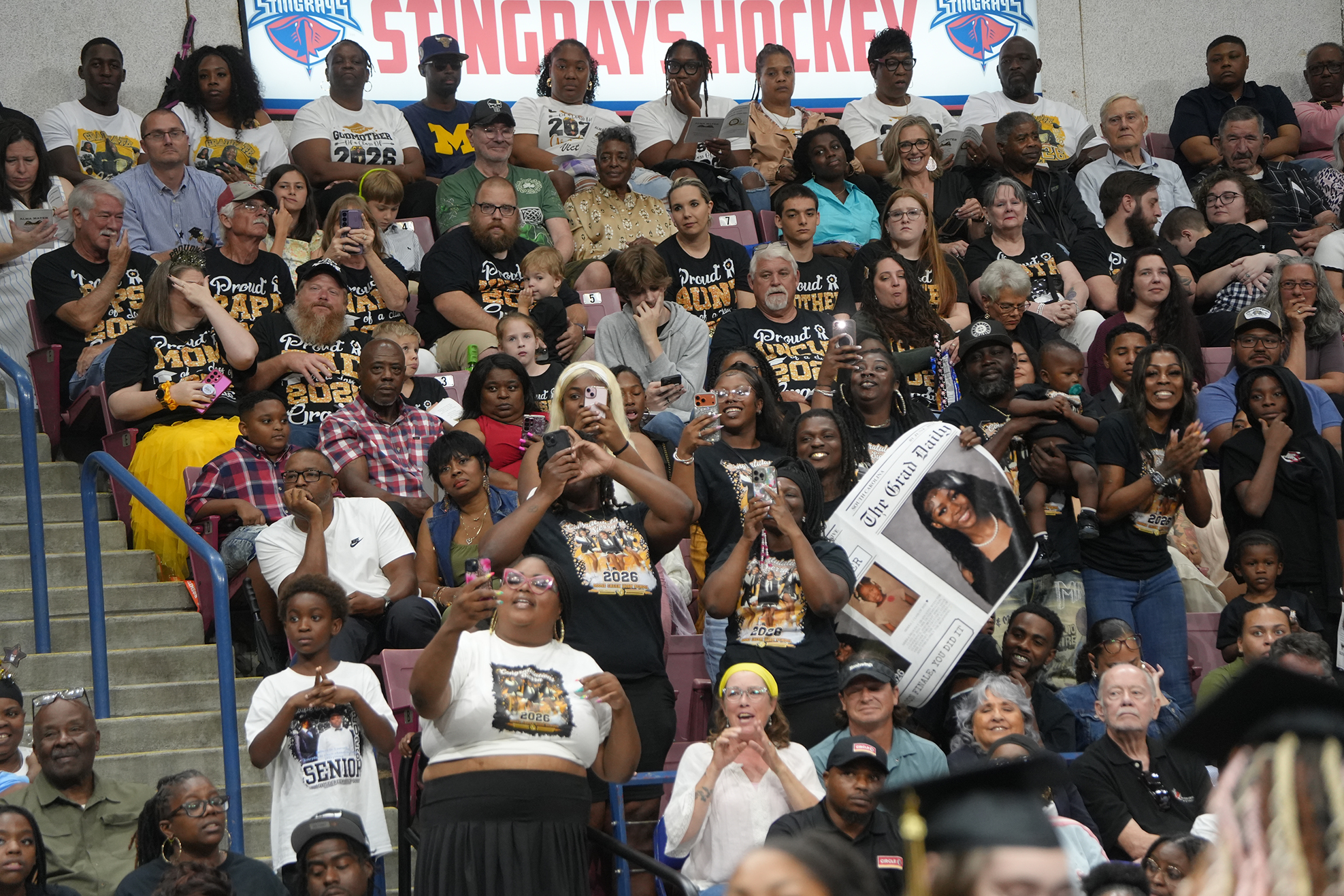 Audience members cheer during a college hockey game, many wearing white T-shirts with colorful designs.