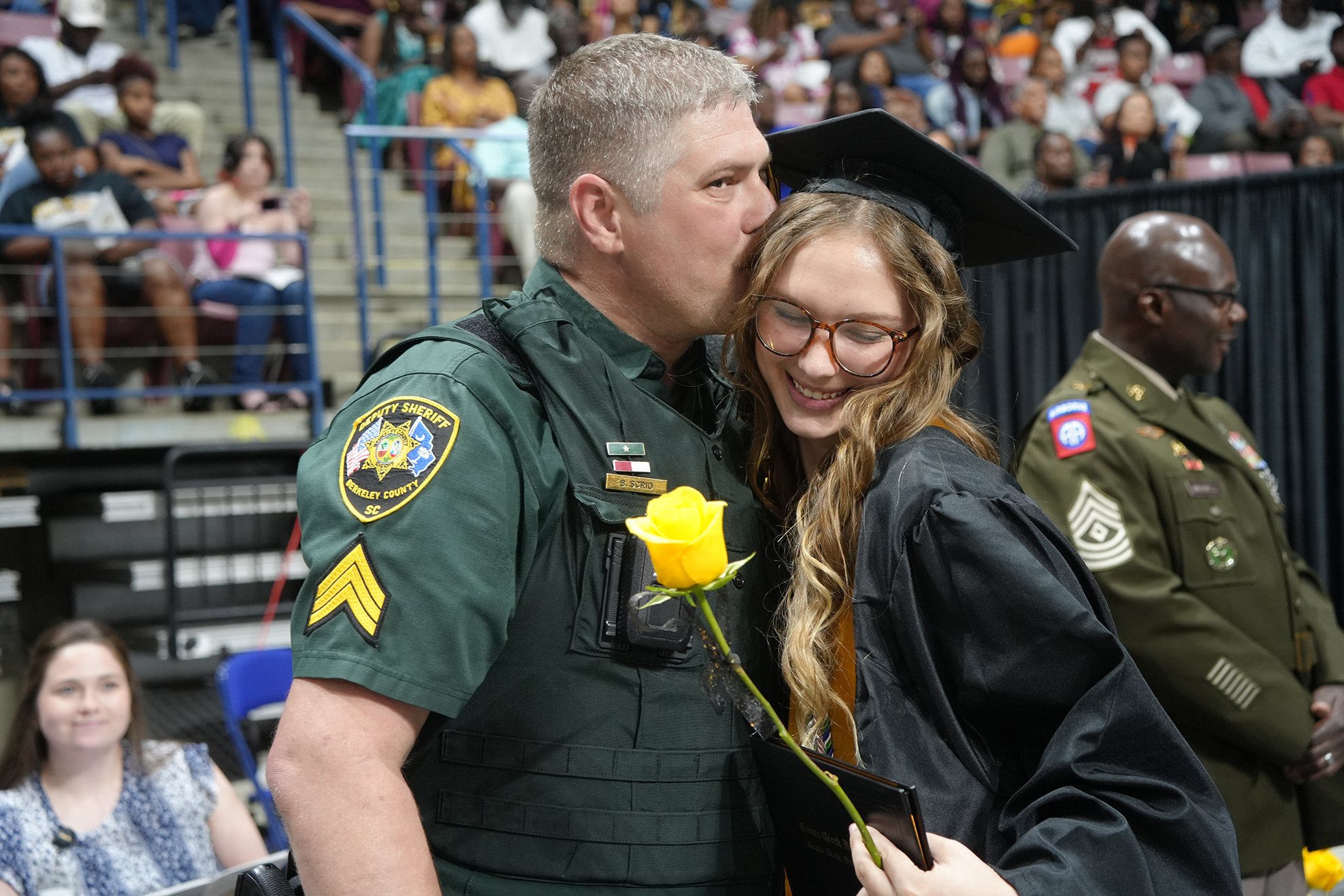 A police officer in uniform kisses a woman in a graduation gown. They hold a yellow rose.