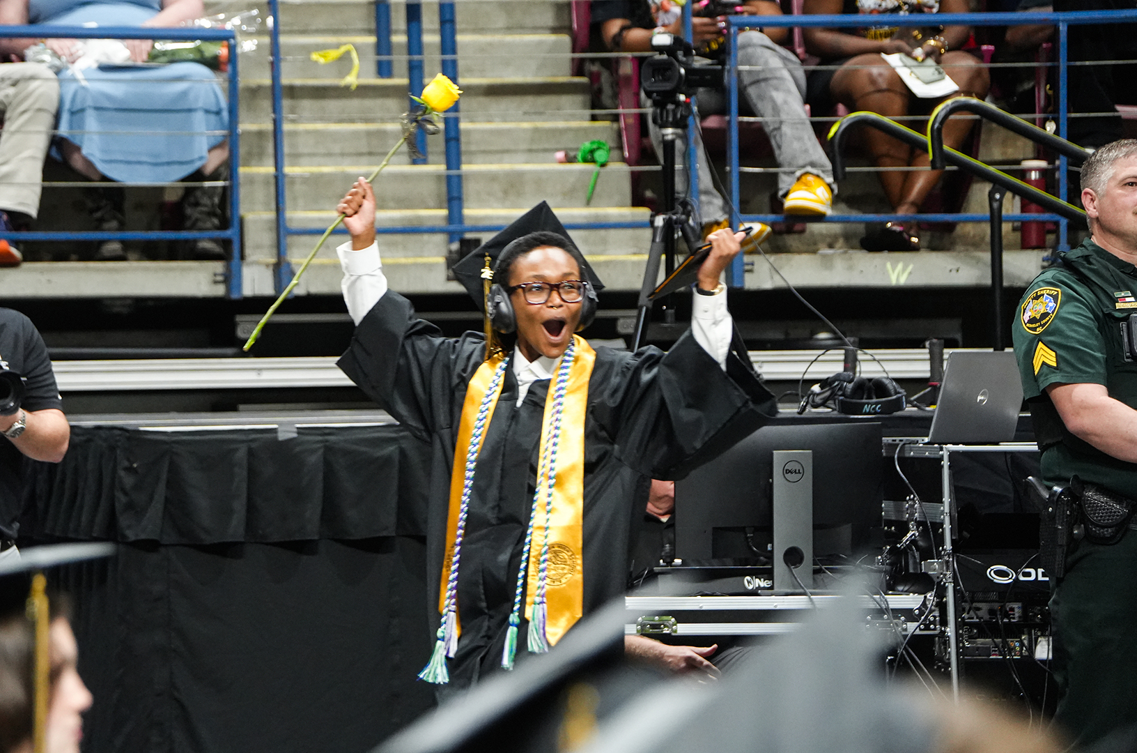 A graduate in a gown and cap holds a flower while raising both arms in a stadium.