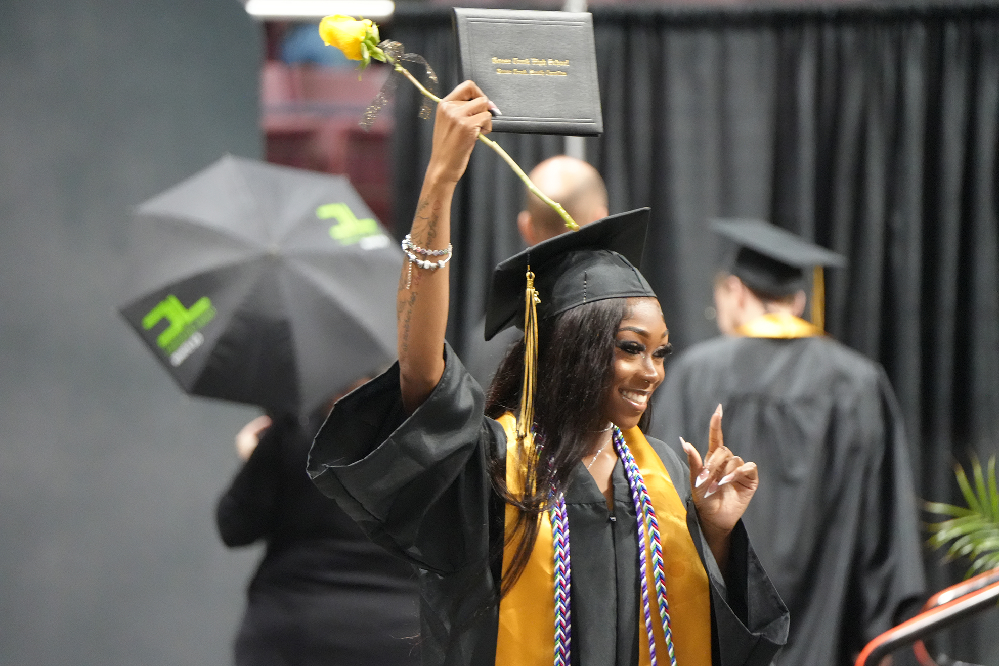 A woman in a black gown with a yellow flower on her cap raises her hand during a graduation.