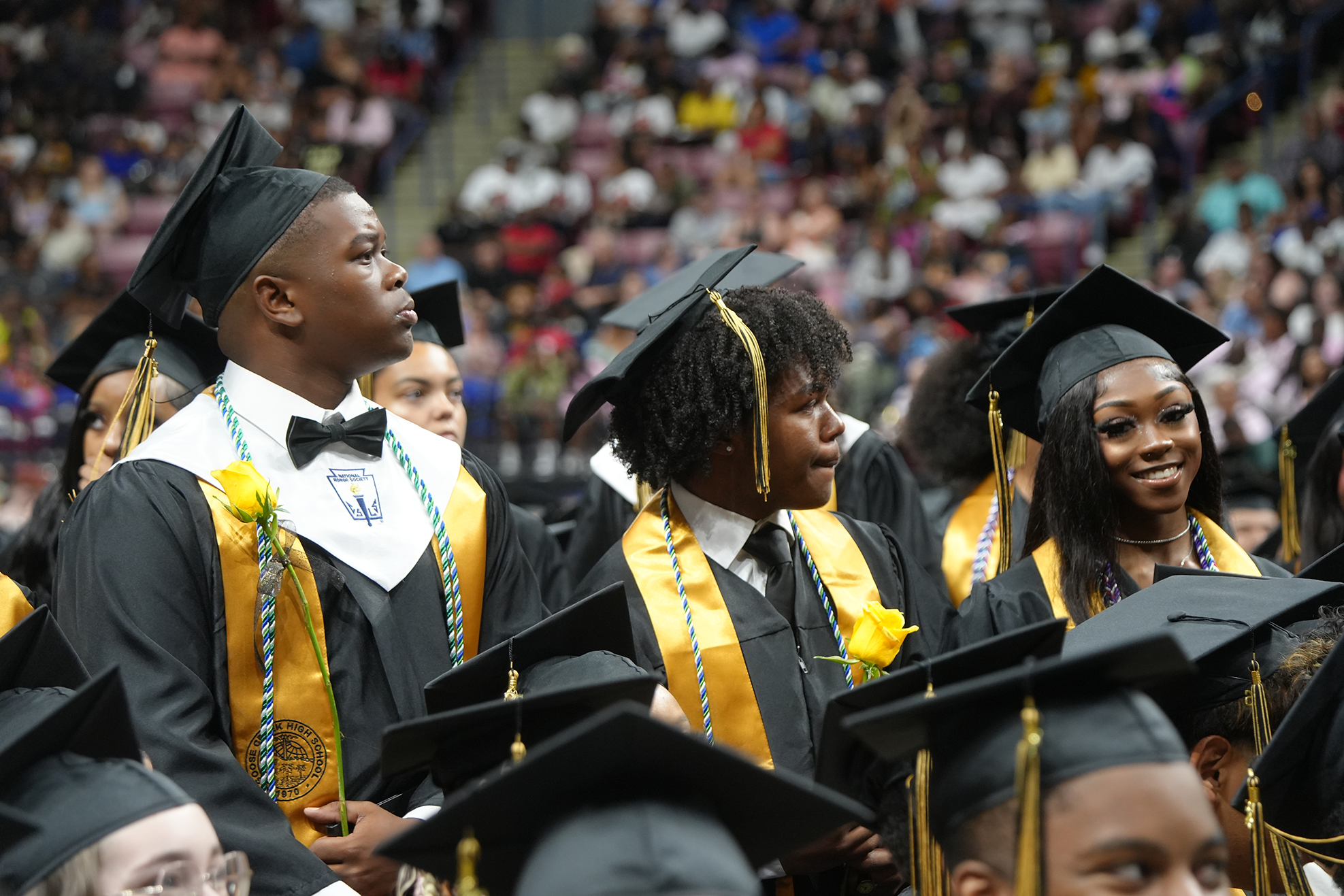 A group of young people in graduation attire stands together, looking up. Behind them, a large audience fills the seats of a stadium.