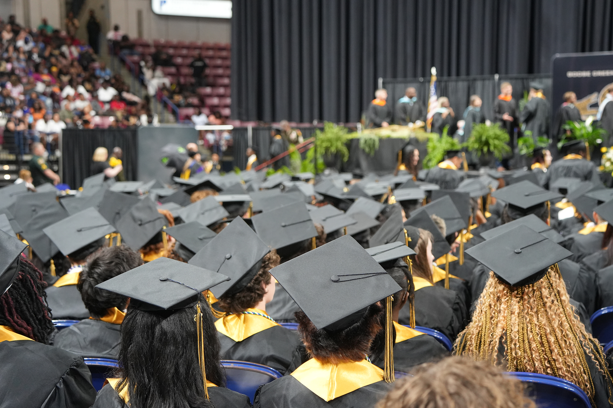 Large audience in a hall, many in black caps and gowns, looking toward a stage with people.