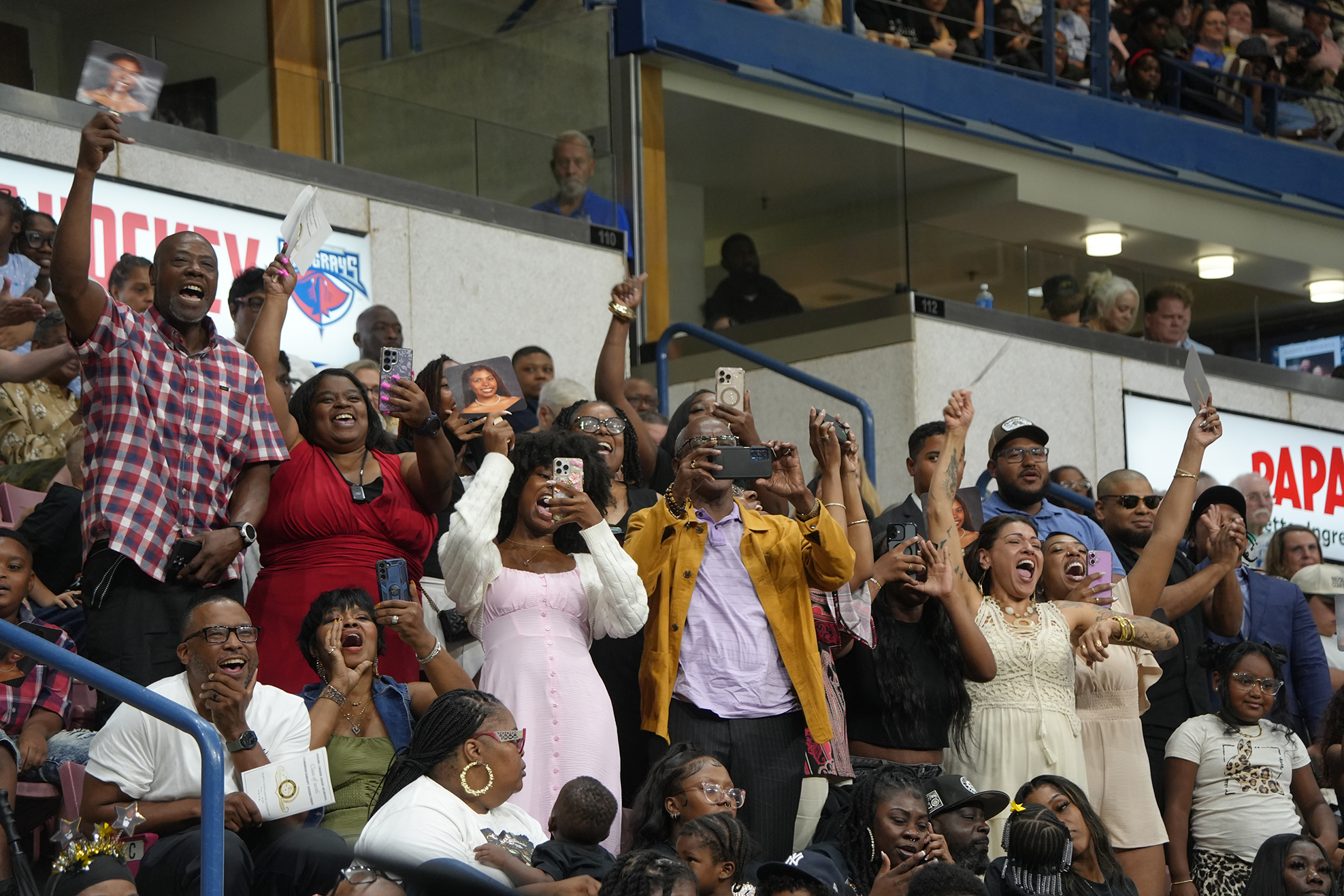 A diverse group of people in a stadium stands, raising hands and holding phones, some wearing glasses.