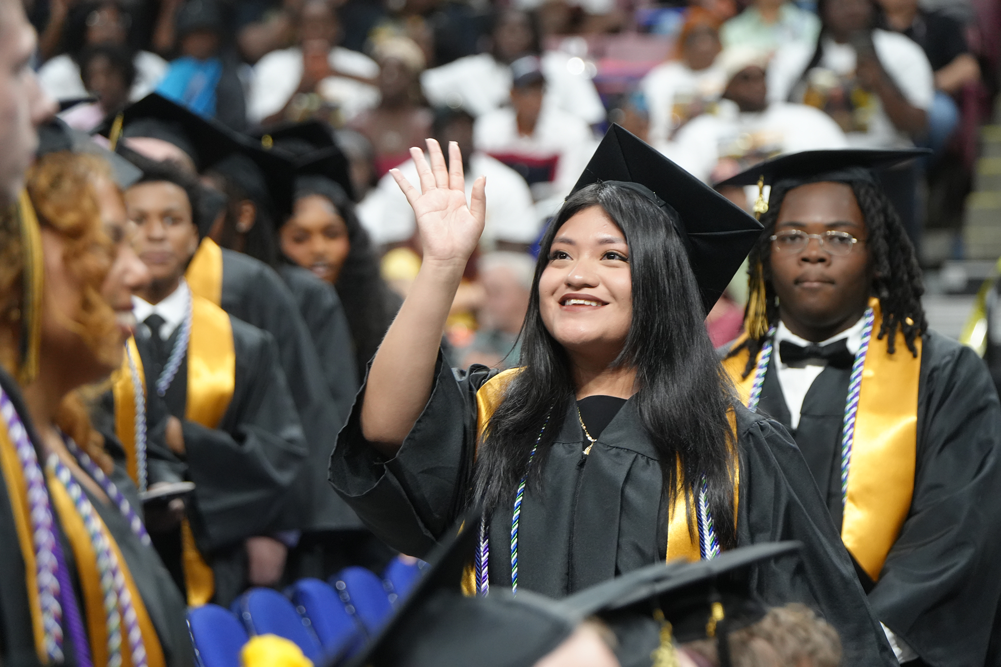 Several people in caps and gowns, one person waves to the crowd, smiling, with a blurred audience in the background.