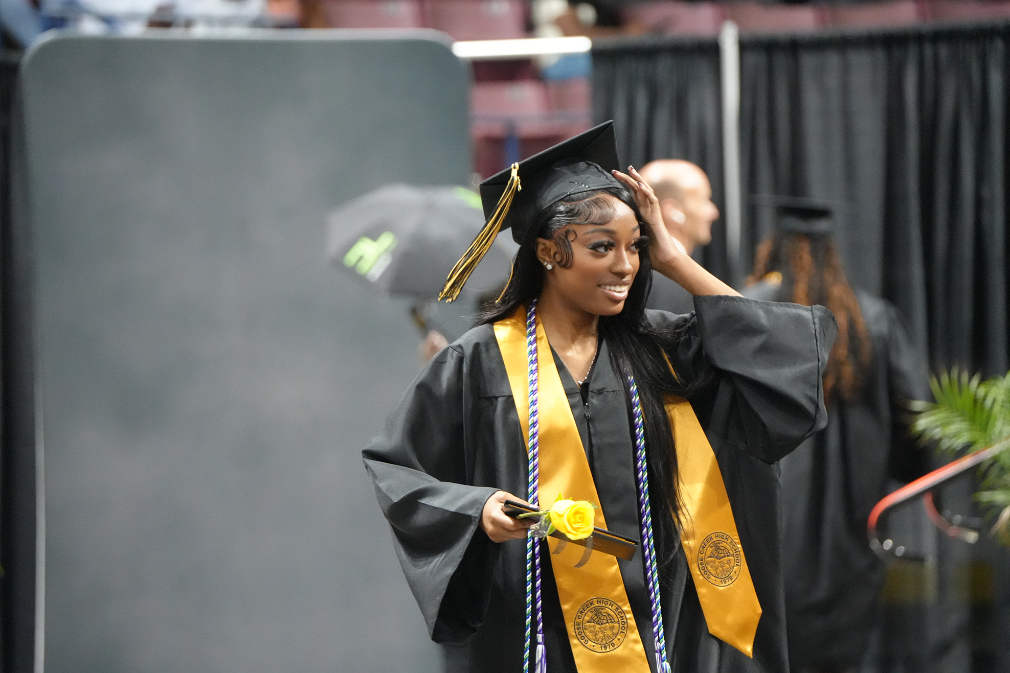A woman in a black graduation gown with a yellow stole adjusts her cap on a stage. Behind her, other graduates walk with umbrellas, and a plant is visible.
