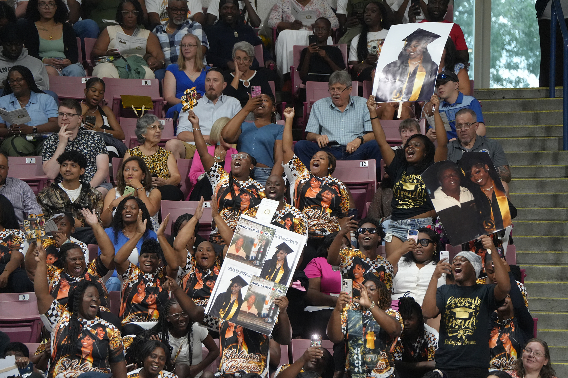 A crowd in a stadium cheering and holding up pictures. Some wear graduation gowns and hats.