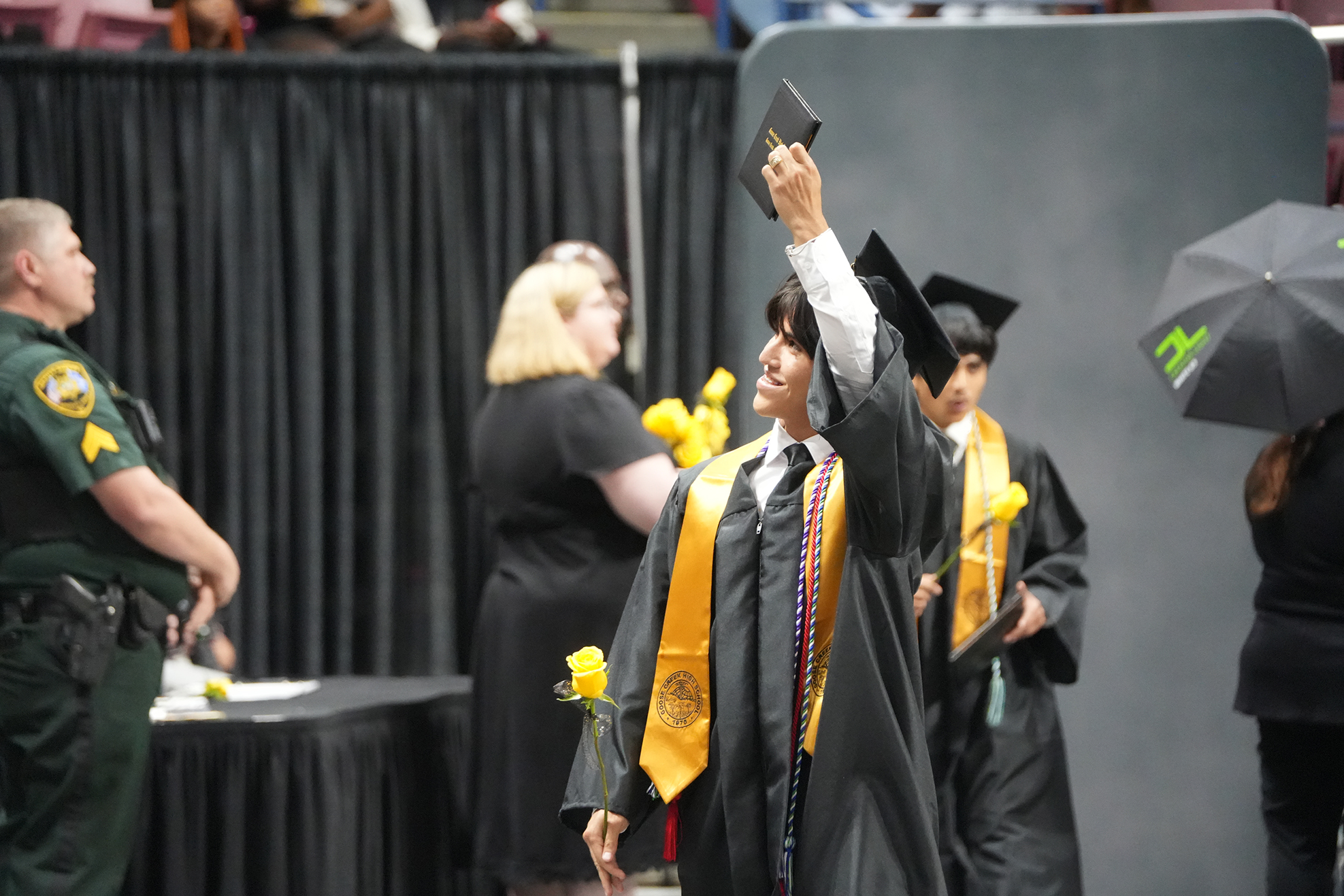 A person in a graduation gown and hat raises their diploma. A police officer stands nearby.