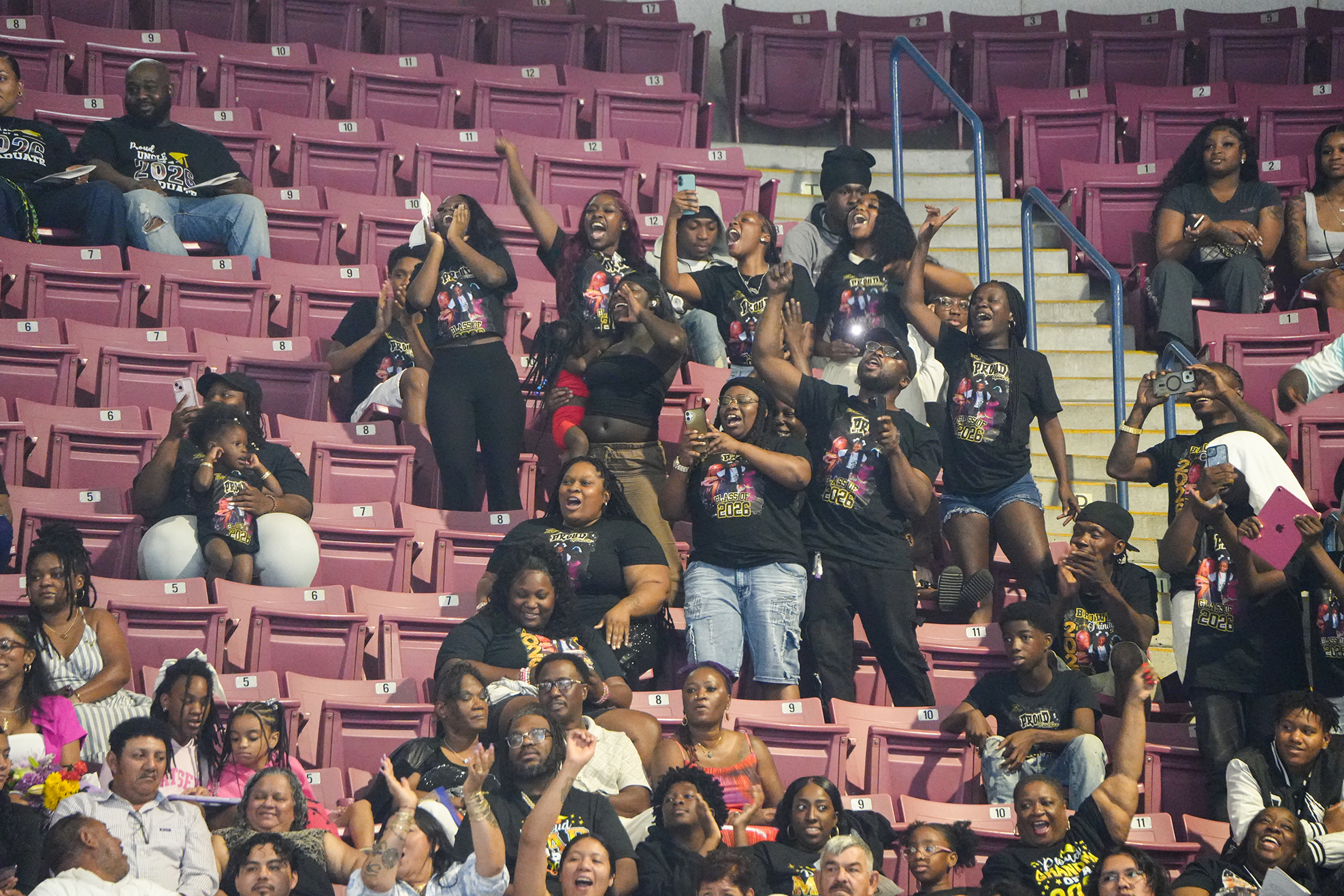Stadium full of people, many wearing black shirts, cheering and standing with hands raised.