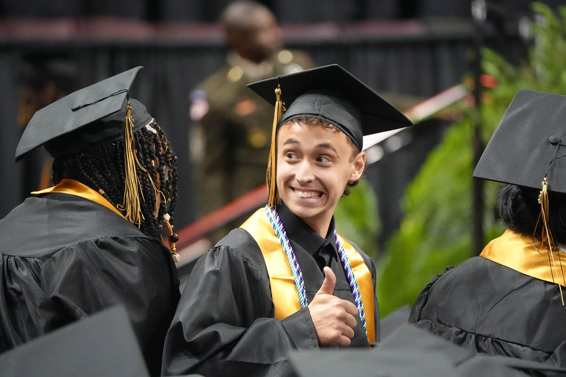 A smiling person in a black graduation gown and cap gives a thumbs up, with blurred students and a background.