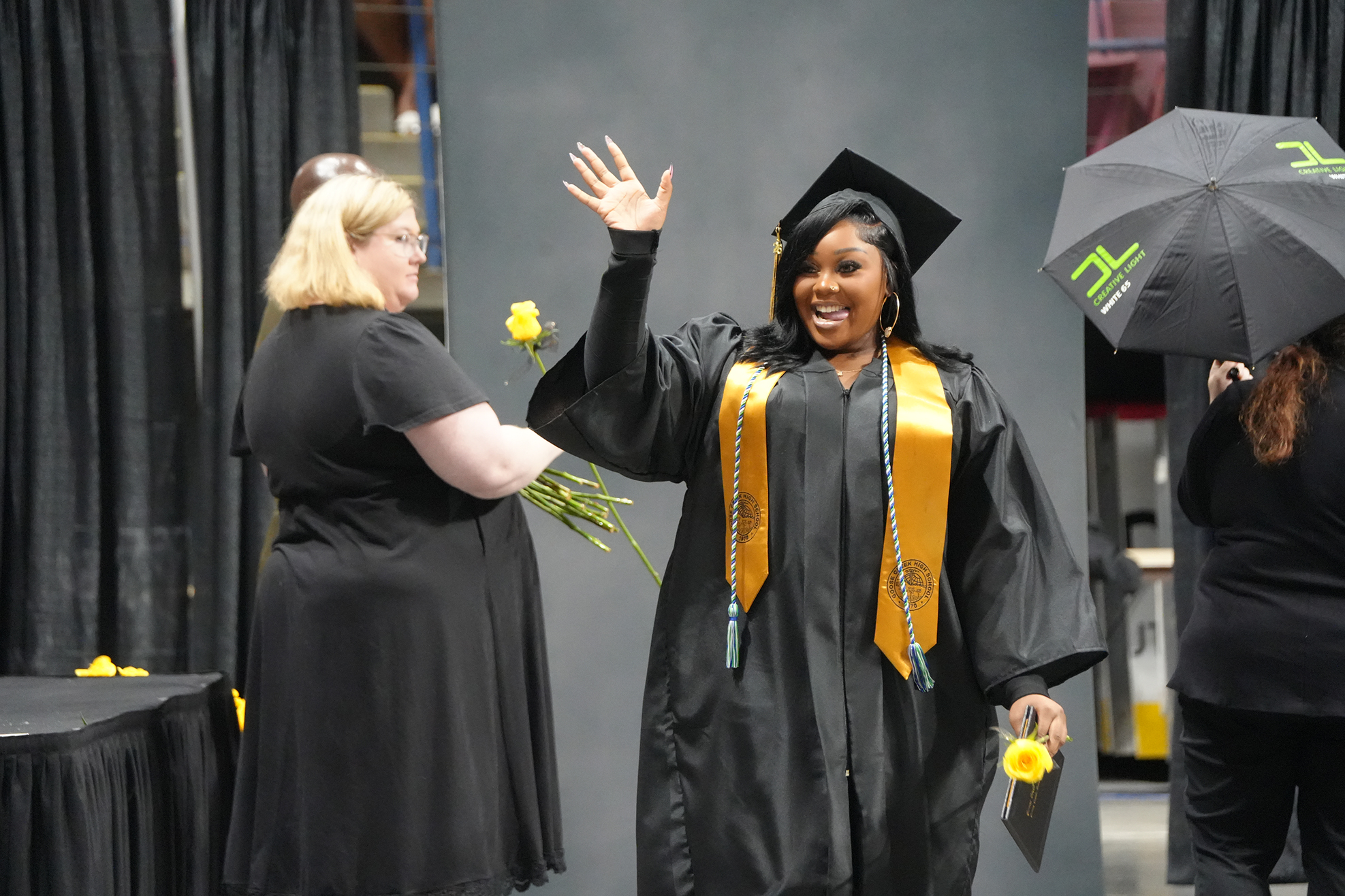 A woman in a black graduation gown waves, another woman holds flowers, and a third holds a black umbrella.