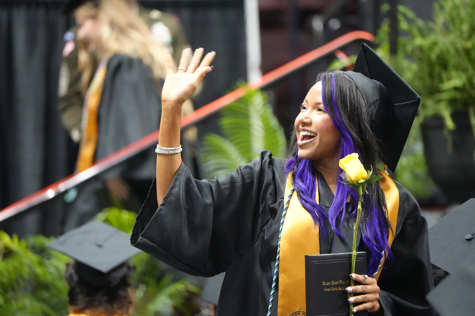 A person with purple hair, waving, and smiling at a graduation ceremony, wearing a cap and gown.