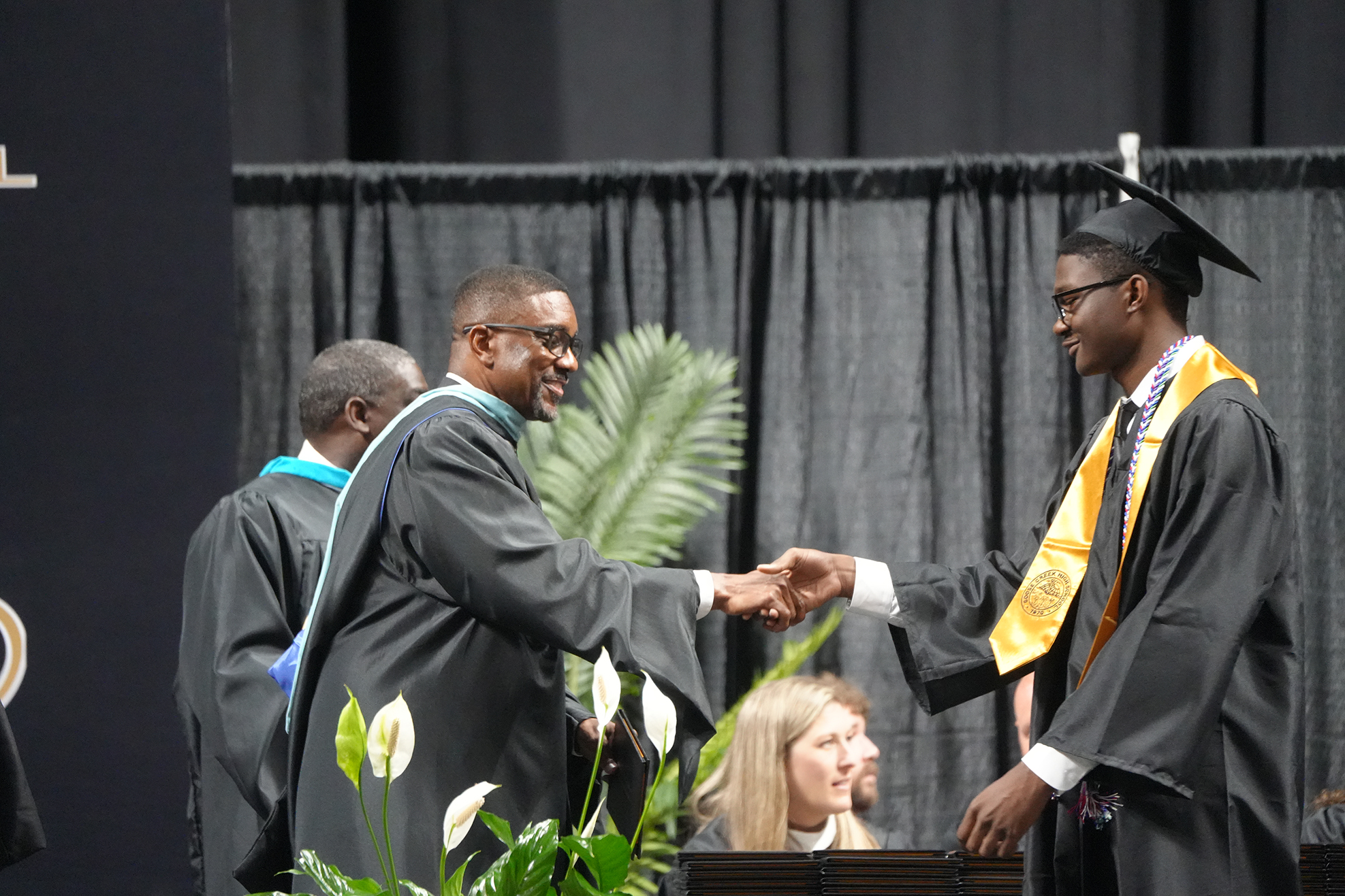 Two men, in caps and gowns, shake hands in front of a stage backdrop. Behind them, a third man stands and two women watch.