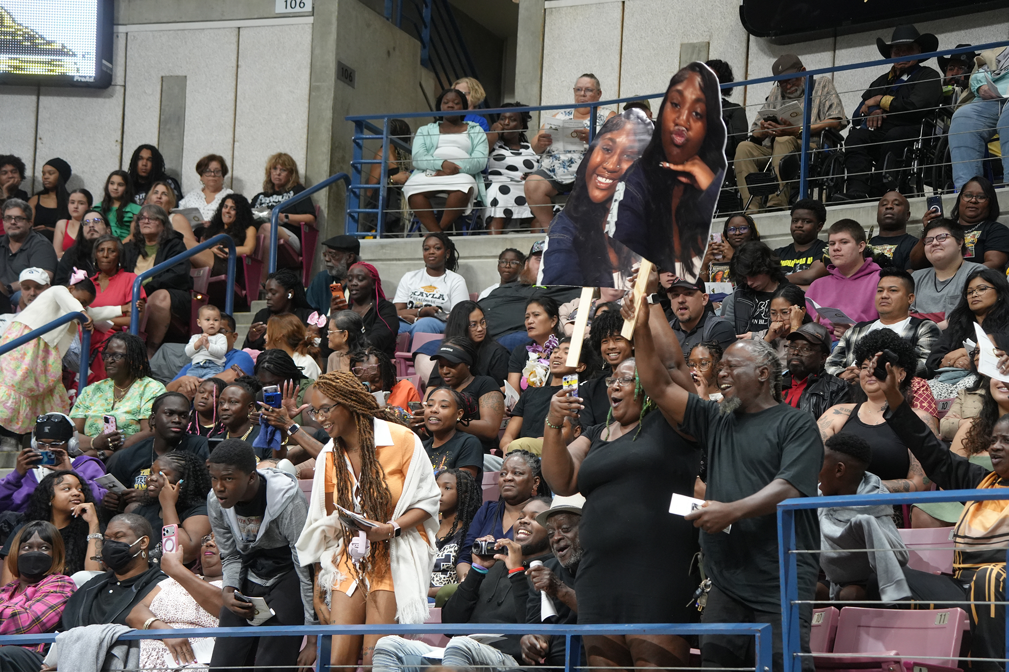 Audience seated in stands, with two people holding up a poster of two women, one in a spotlight.