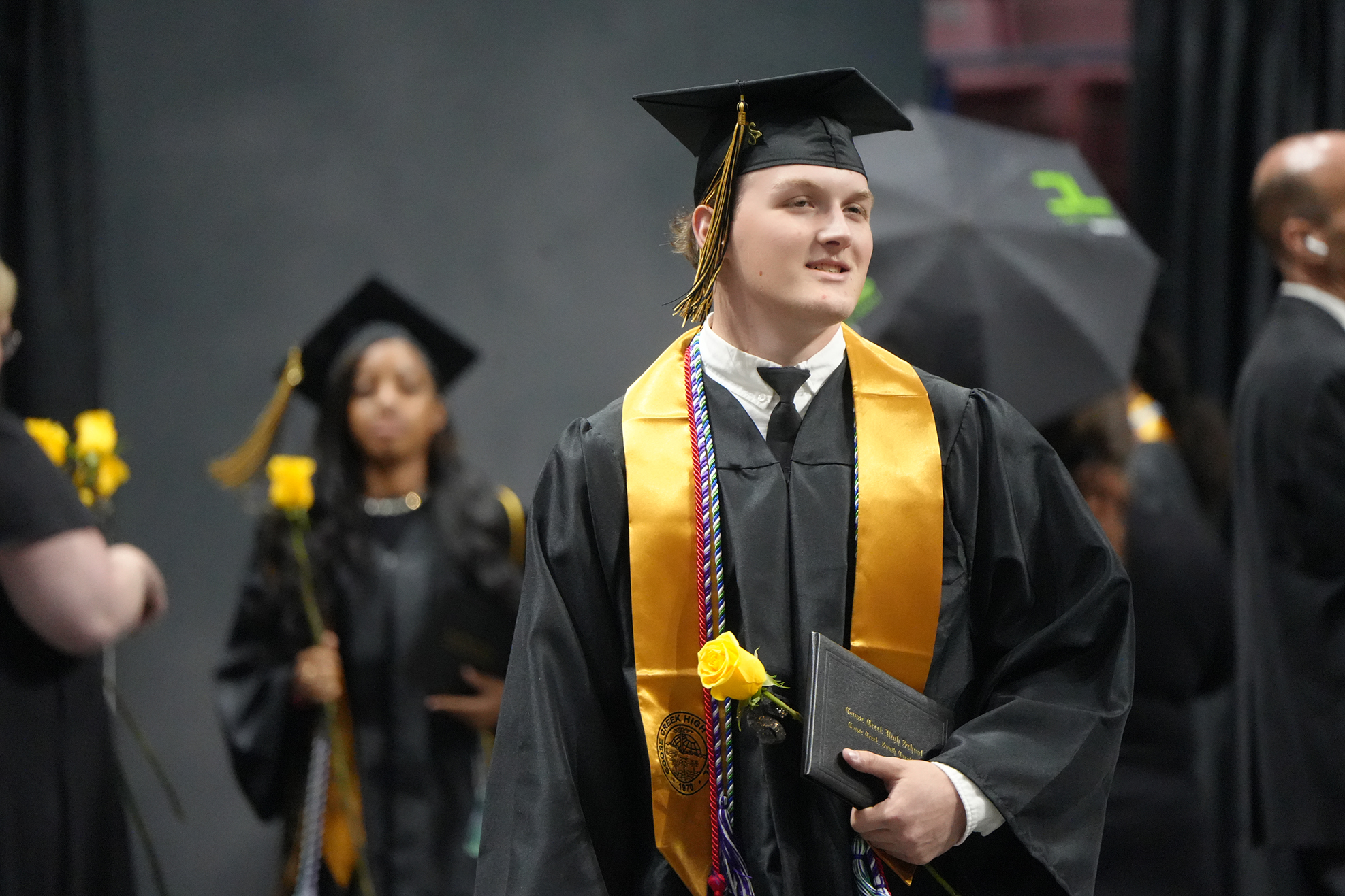 A person in a black graduation gown with a yellow sash holds a diploma and a yellow rose.