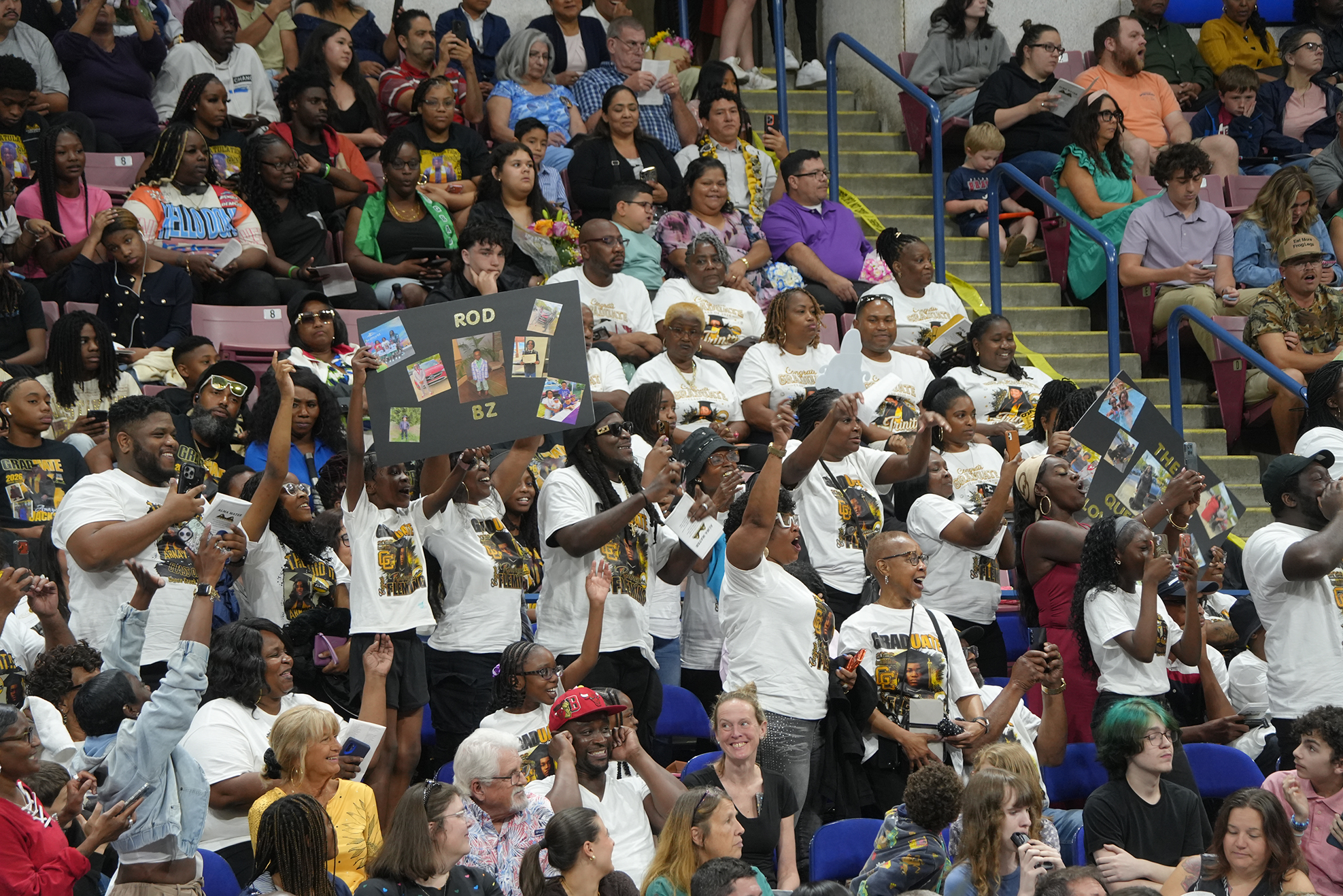 Diverse group of people in a stadium, some in white shirts, holding signs and clapping.