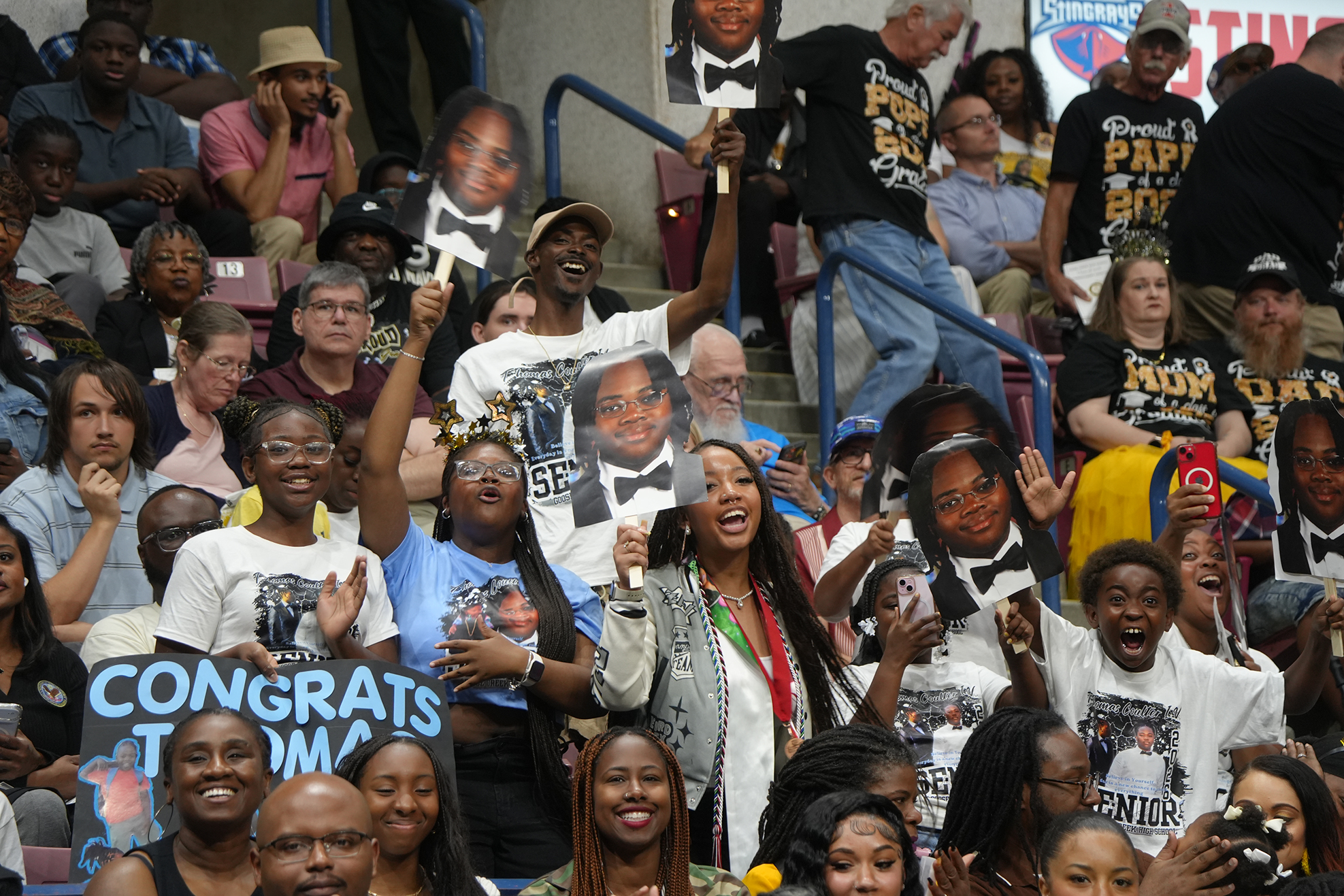 A crowd of people in an auditorium holds up signs and cutouts of people, some cheering.