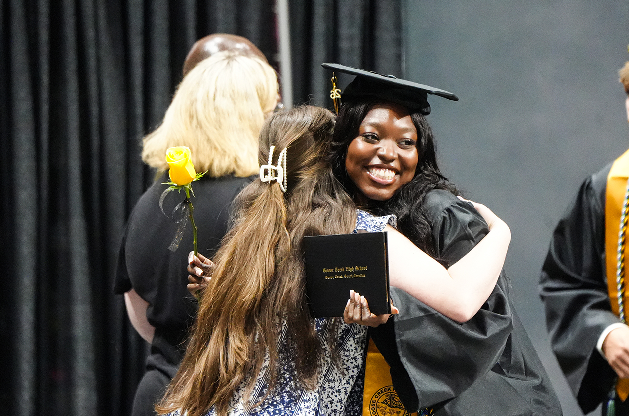 Two women in academic robes, one holding a diploma, hugging and smiling on stage. A woman with blonde hair holds a yellow rose.