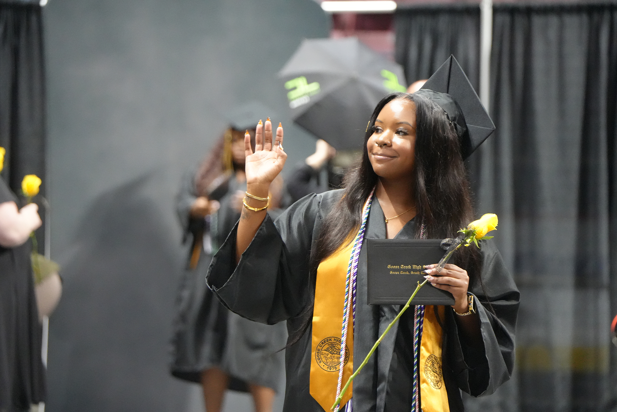 A woman in a graduation gown waves as she holds a diploma and a yellow flower.