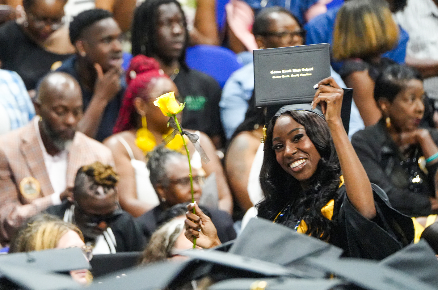 Graduates in black gowns hold up certificates. A graduate holds up a yellow rose. Audience seated behind.