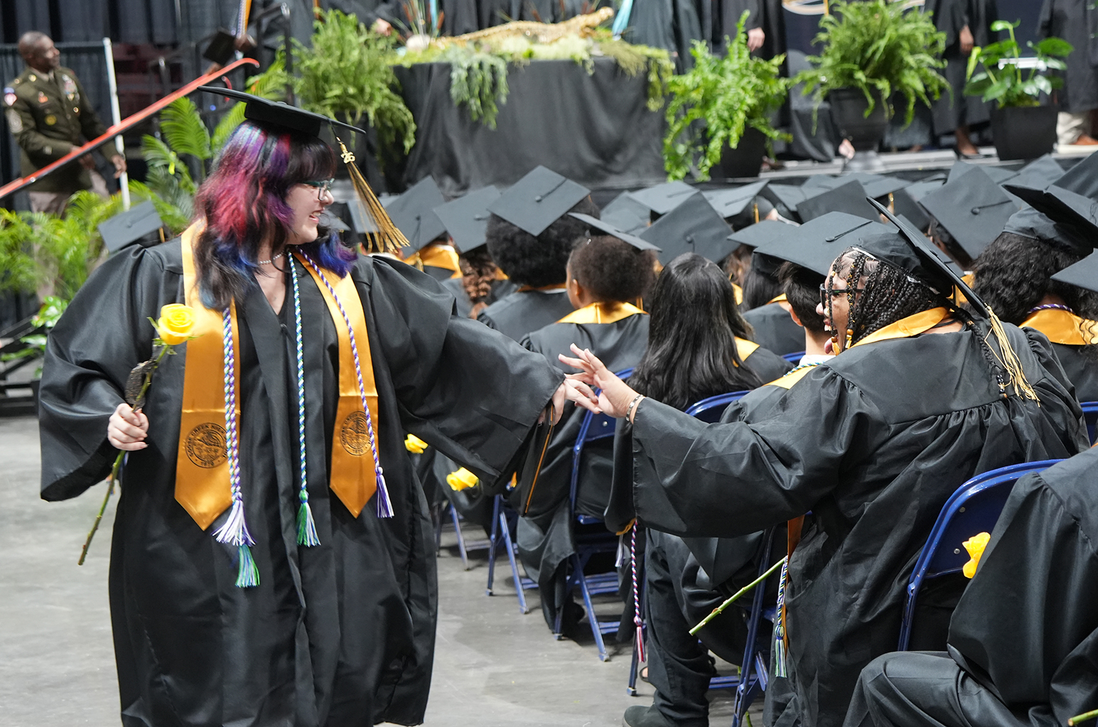 Graduates in black gowns and caps stand in a hall, one extending a hand to another. Potted plants are on the stage behind them.