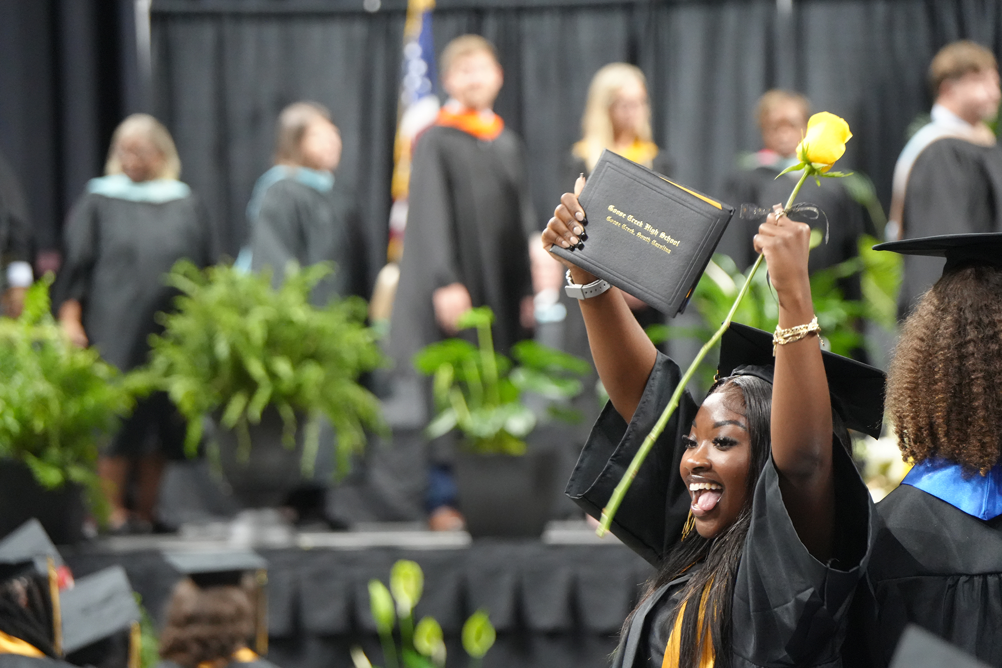 A woman in a black cap and gown holds up a diploma and a yellow rose.