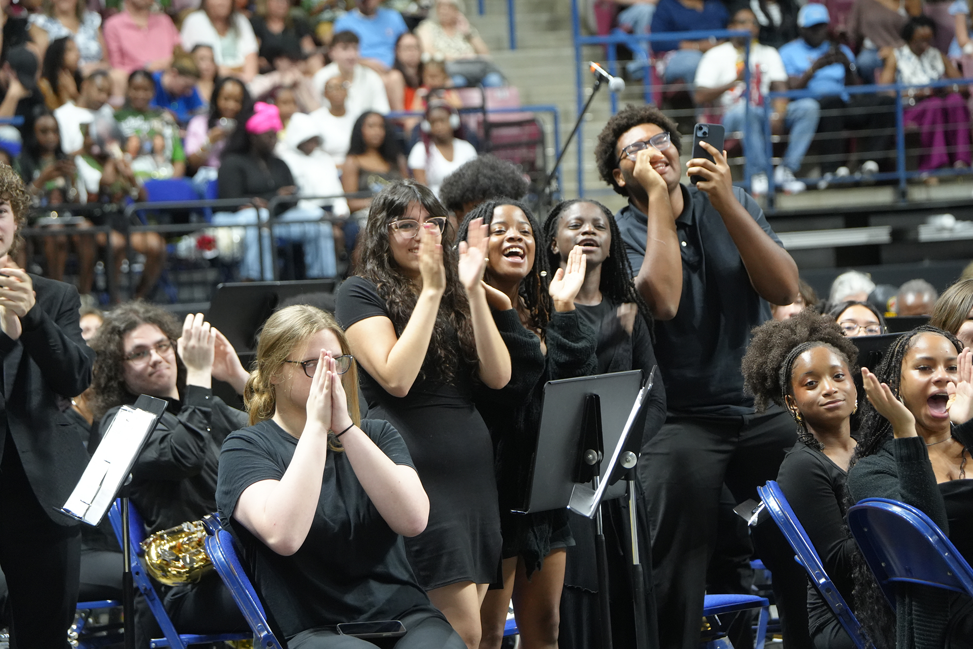 A group of people in black outfits clapping during a performance, seated in front of a stage with a crowd in the background.
