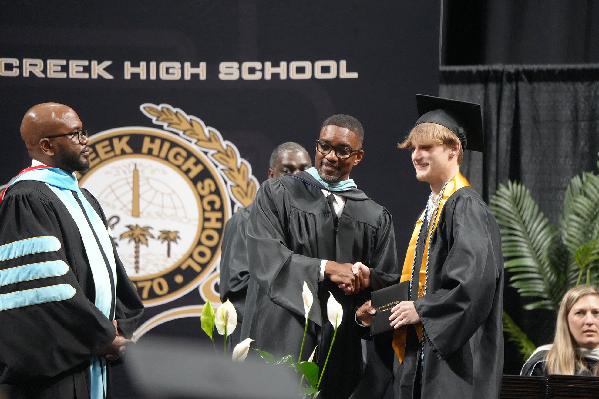 Three people in graduation attire at an event. The central person holds a diploma. Behind them is a stage with plants and a banner reading "Creek High School."