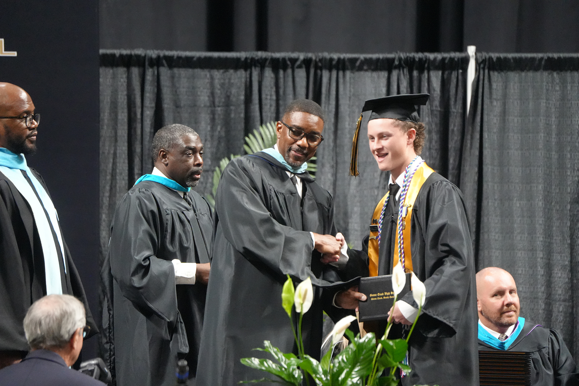 Four men in academic dress on a stage; the man in front exchanges a diploma with a smiling recipient.