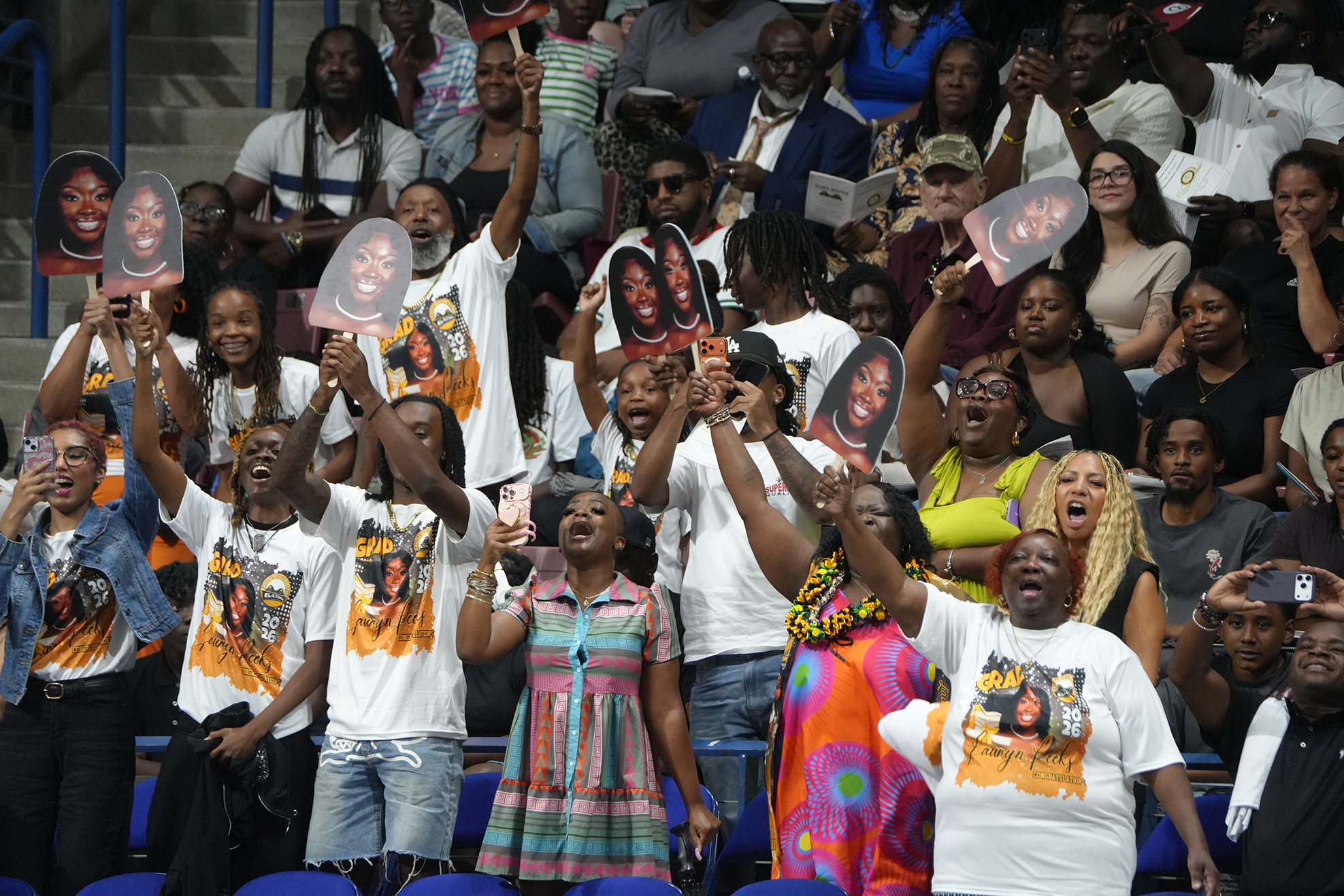 A group of people in a stadium, some holding up signs, others raising their arms, cheering at an event.