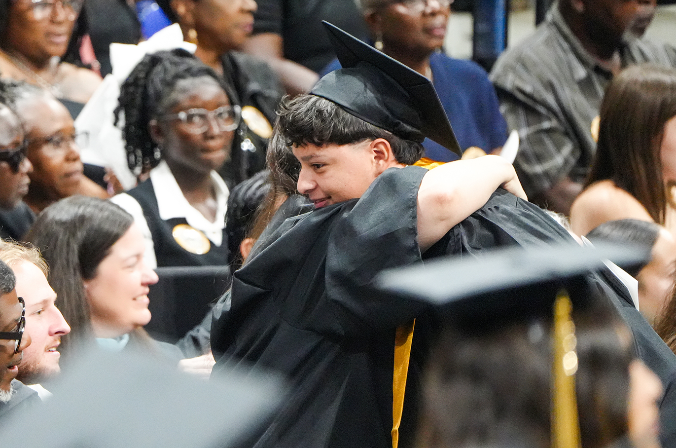 People in a crowded room wear caps and gowns, likely at a graduation ceremony, with two people hugging in the foreground.