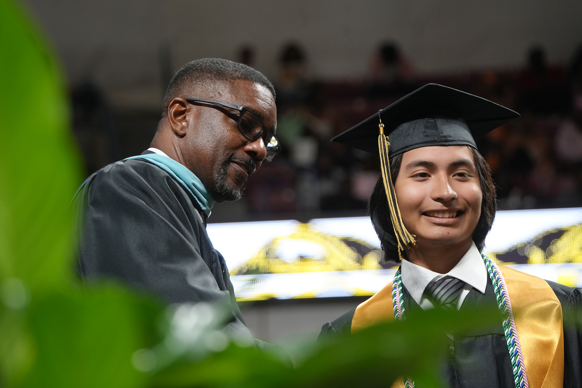 A man and woman in academic regalia at a graduation. The man smiles, and the woman looks on.
