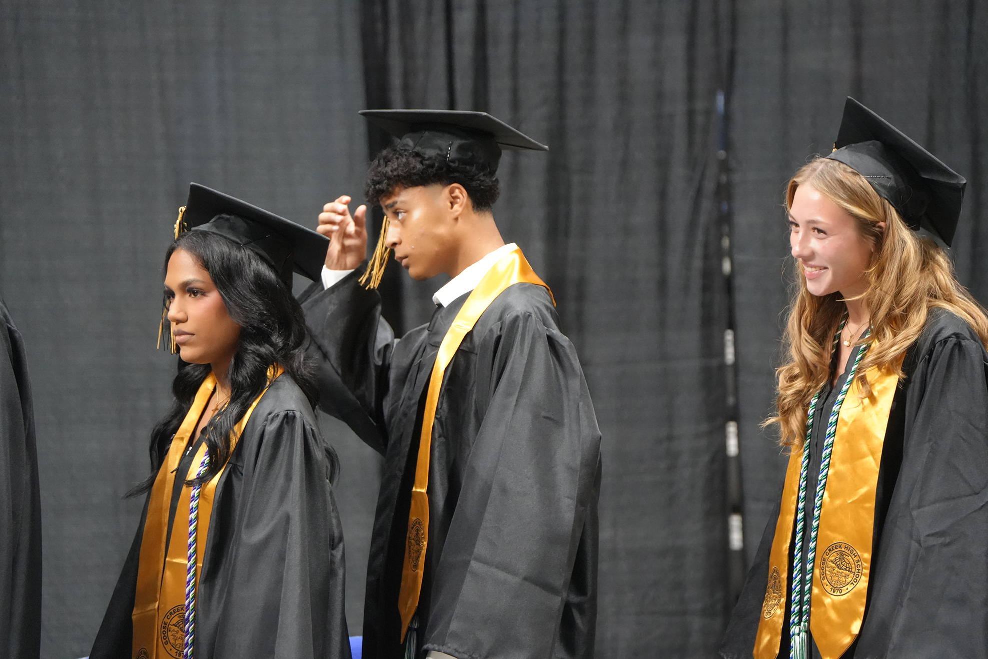 Three individuals in graduation attire, each wearing a cap, stand together. One person adjusts another's cap. The background is a solid black curtain.