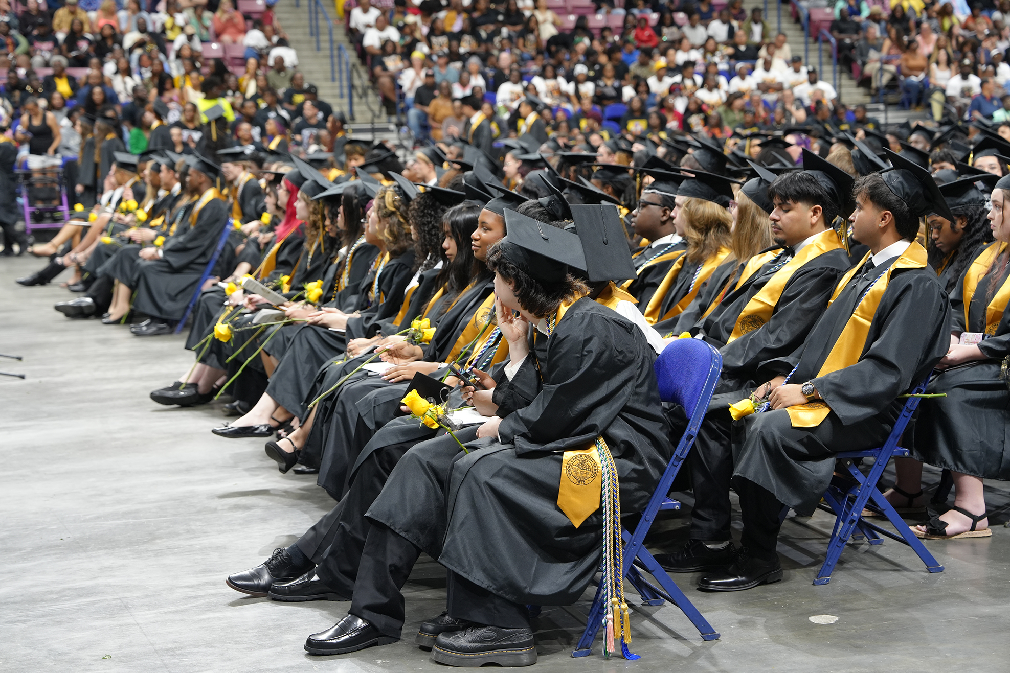 Graduates in black gowns and caps sit in rows of blue chairs in a large auditorium.