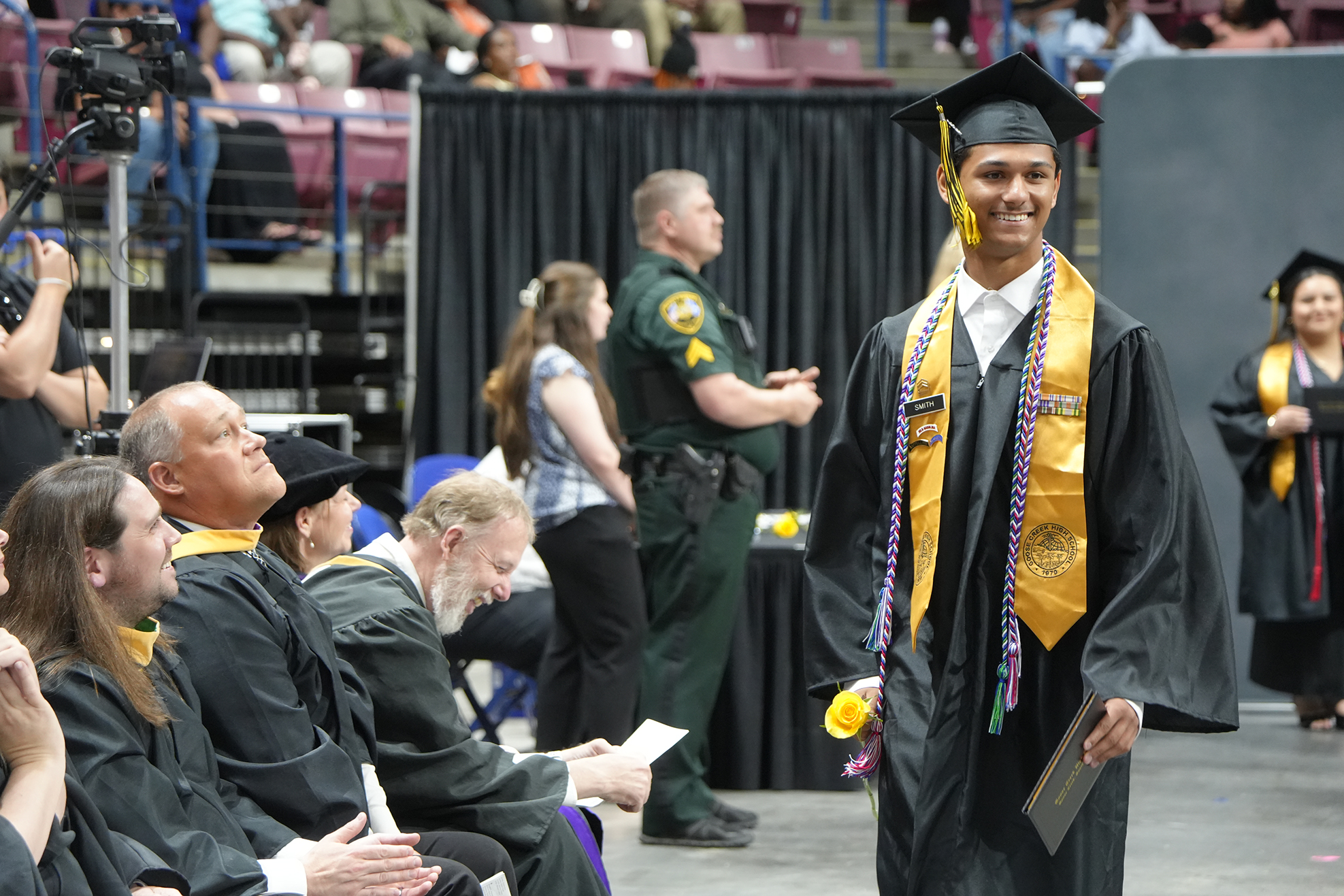 A graduate in a black gown and yellow stole walks across a stage at a ceremony. People sit in the audience and watch.