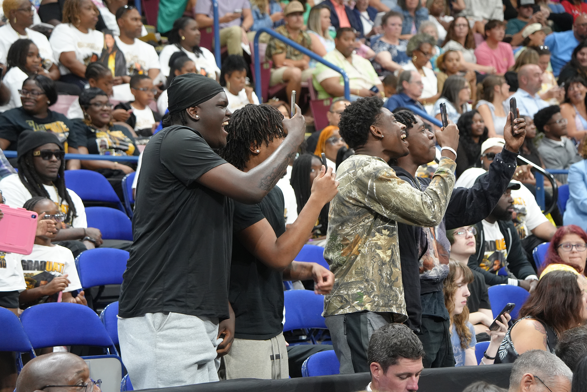 Three men, one singing into a microphone, stand in a large audience with blue chairs.