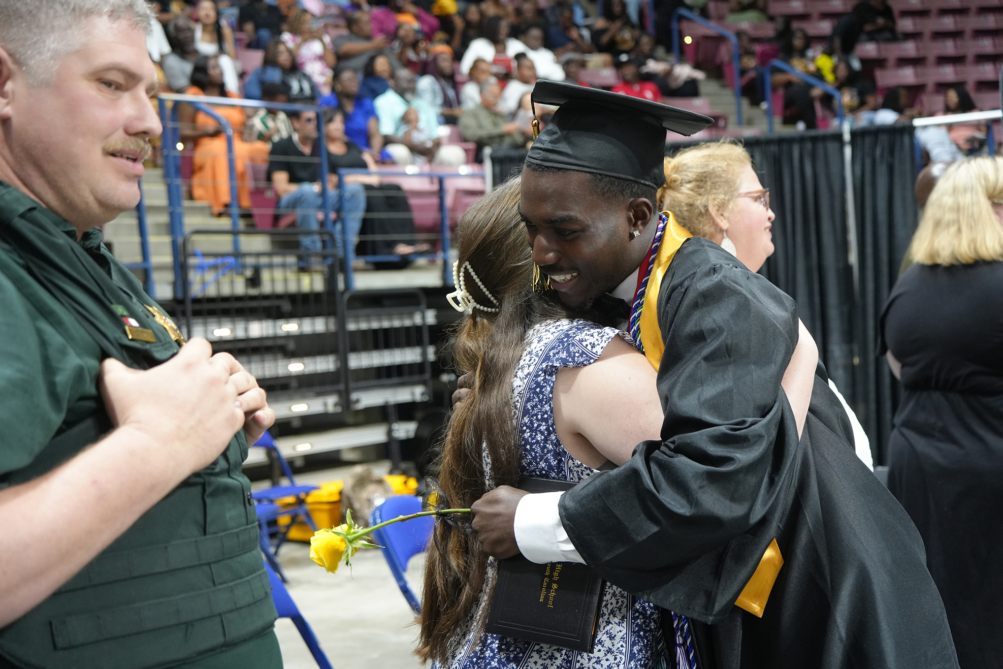 A man in a cap and gown hugs a woman during a graduation ceremony. Spectators fill the bleachers in the background.