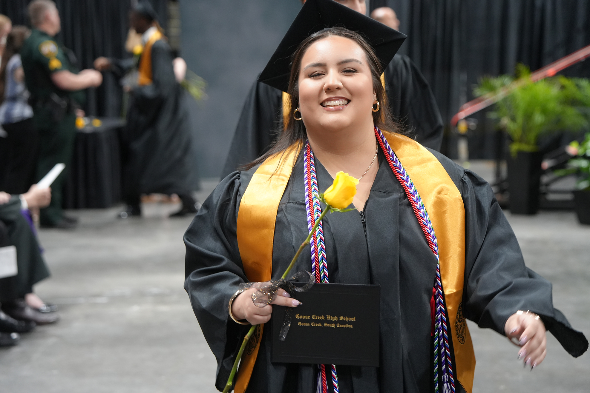 A smiling graduate in a black gown and mortarboard, holding a yellow flower, walks in a corridor.