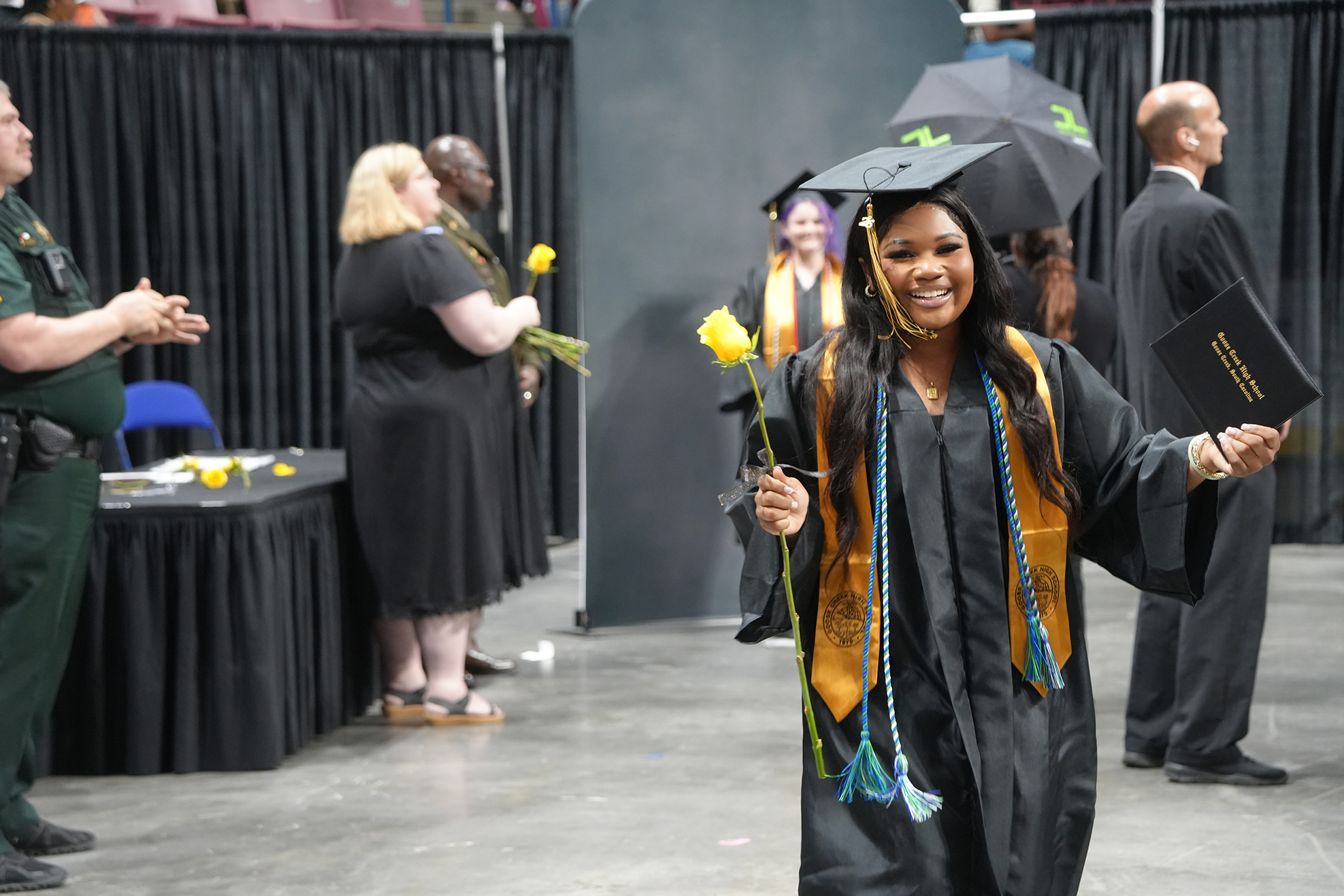 A woman in a graduation gown holds a yellow flower and a diploma. Behind her, people in black gowns and hats, and others stand.