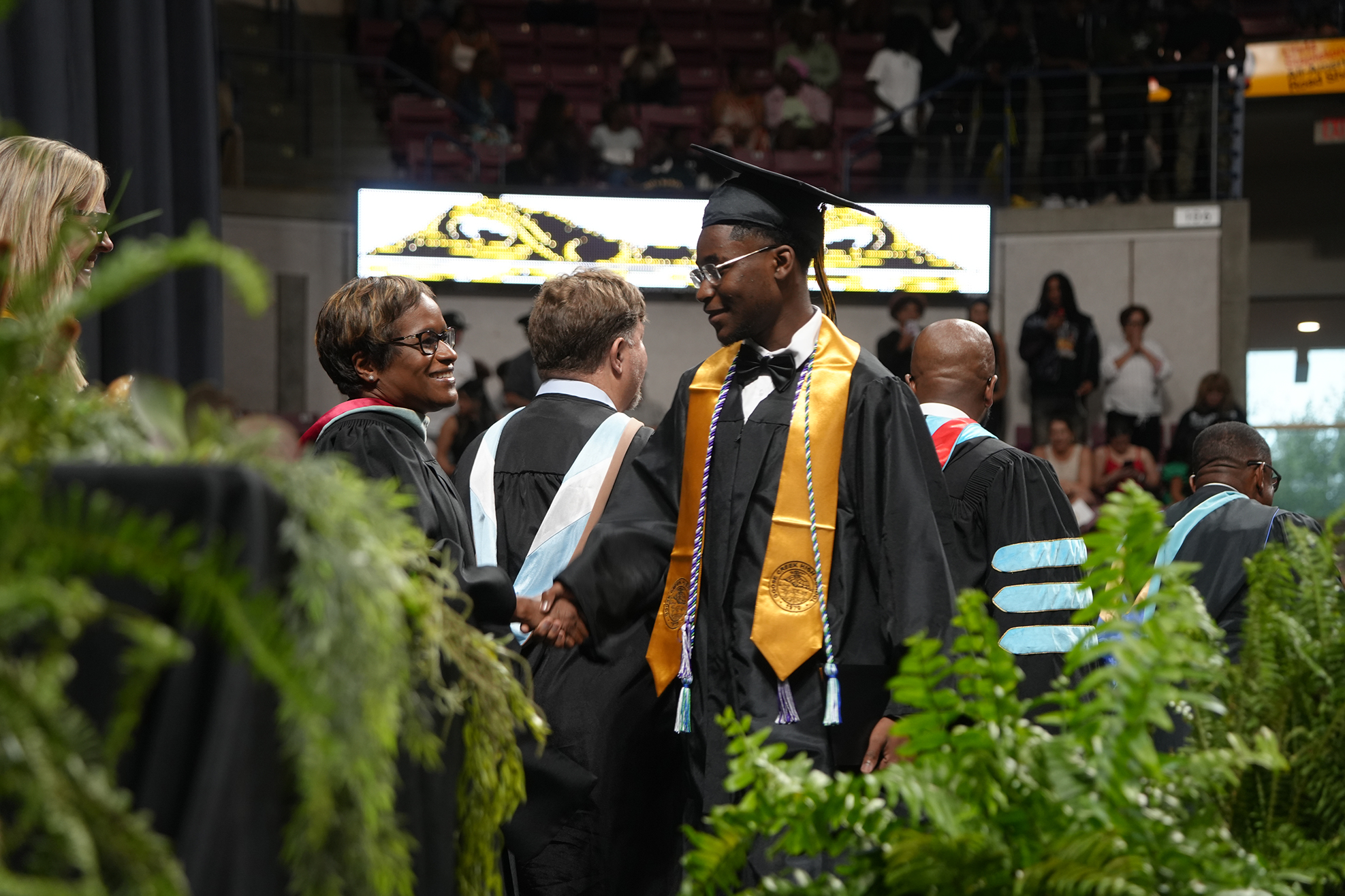 A graduate in a cap and gown shakes hands with a person at a ceremony. Other graduates stand nearby.