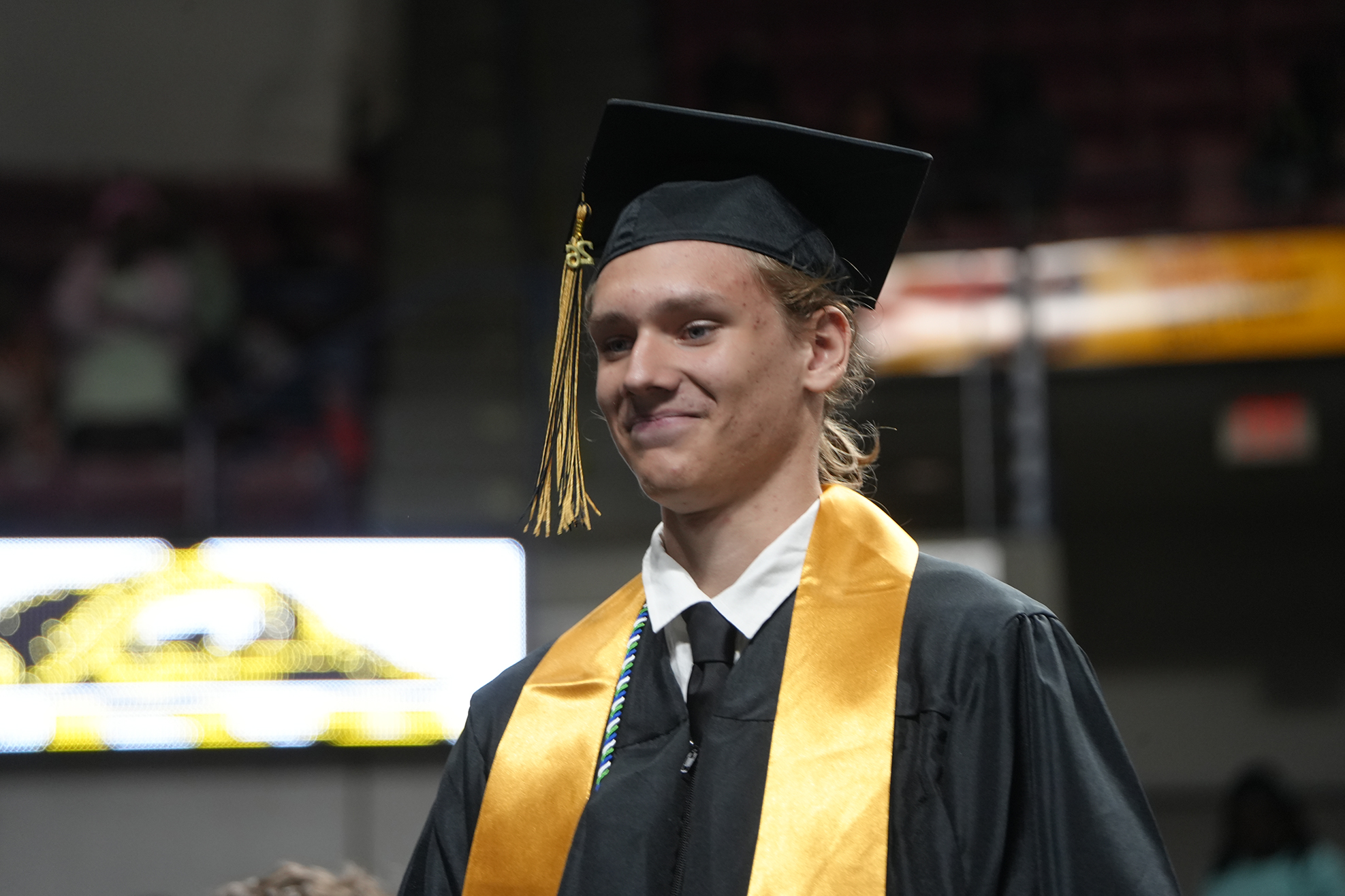 Person in black graduation gown and hat, smiling, stands with yellow sash. Behind them, a large screen shows a graphic.